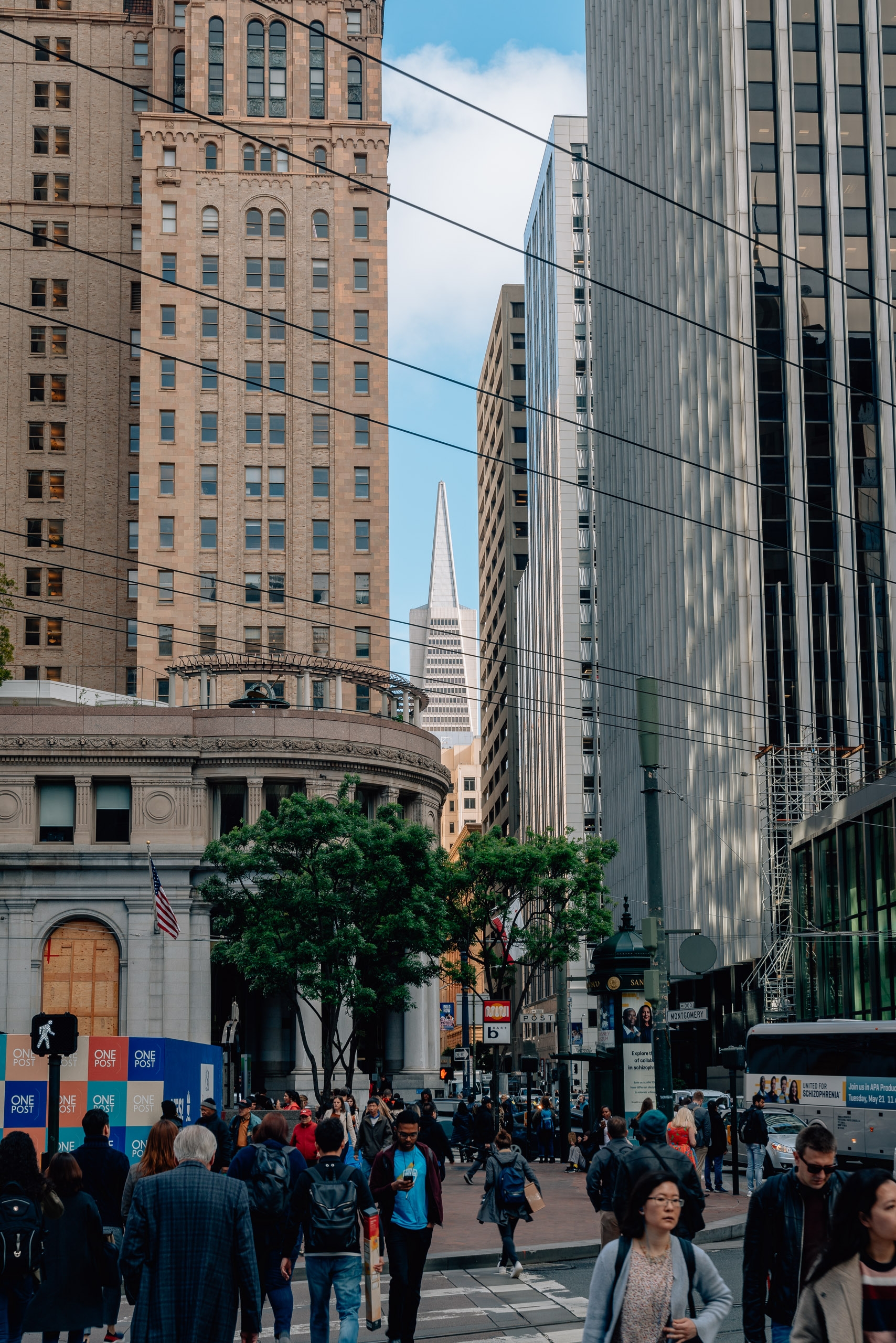 Crowded Market Street pedestrians gather near a classical building framed by towering brick and glass skyscrapers, with the Transamerica Pyramid visible in the distance under clear blue sky