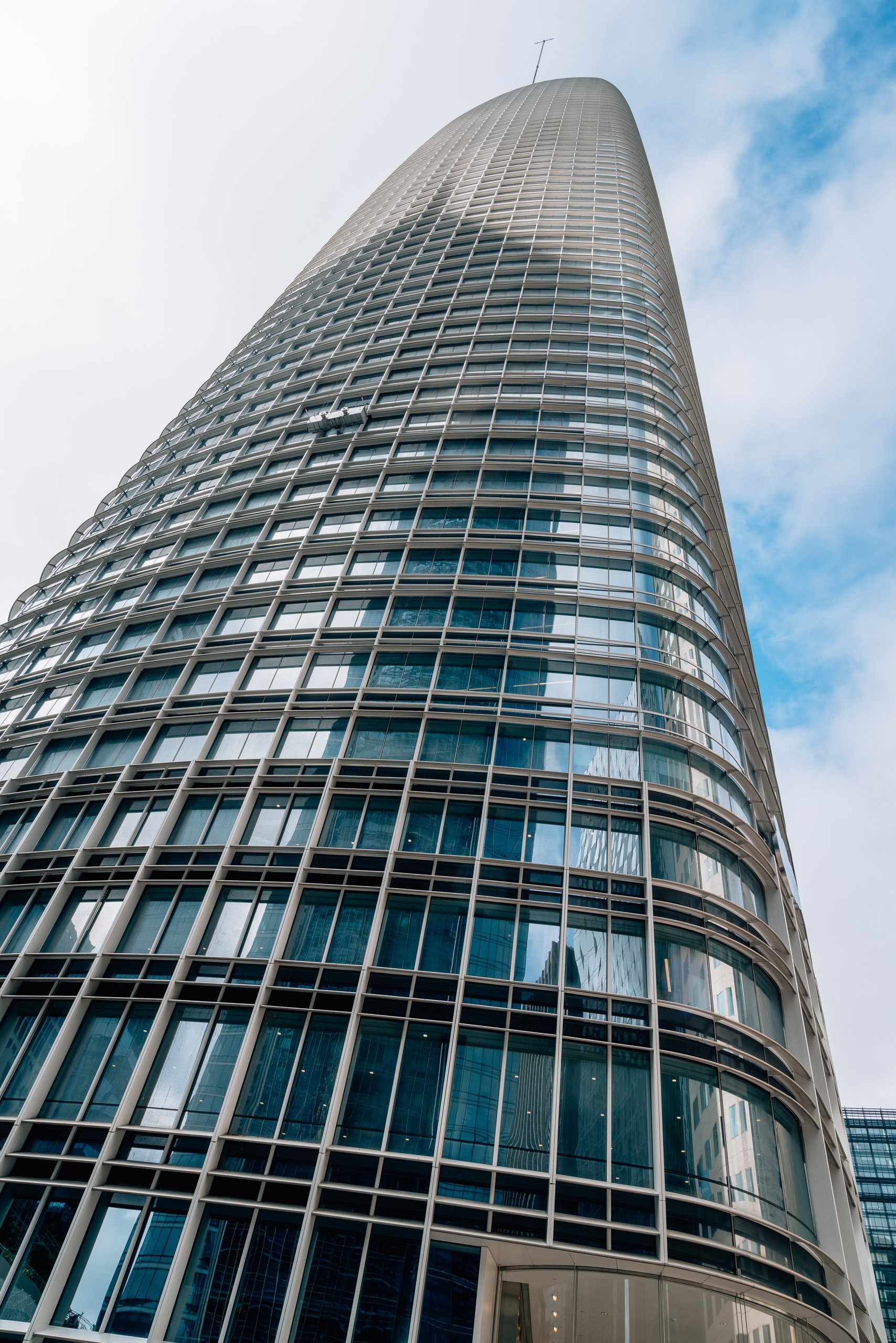 A cylindrical glass and steel skyscraper tapers upward against a partly cloudy sky, its gridded facade reflecting sunlight and blue tones from the surrounding environment