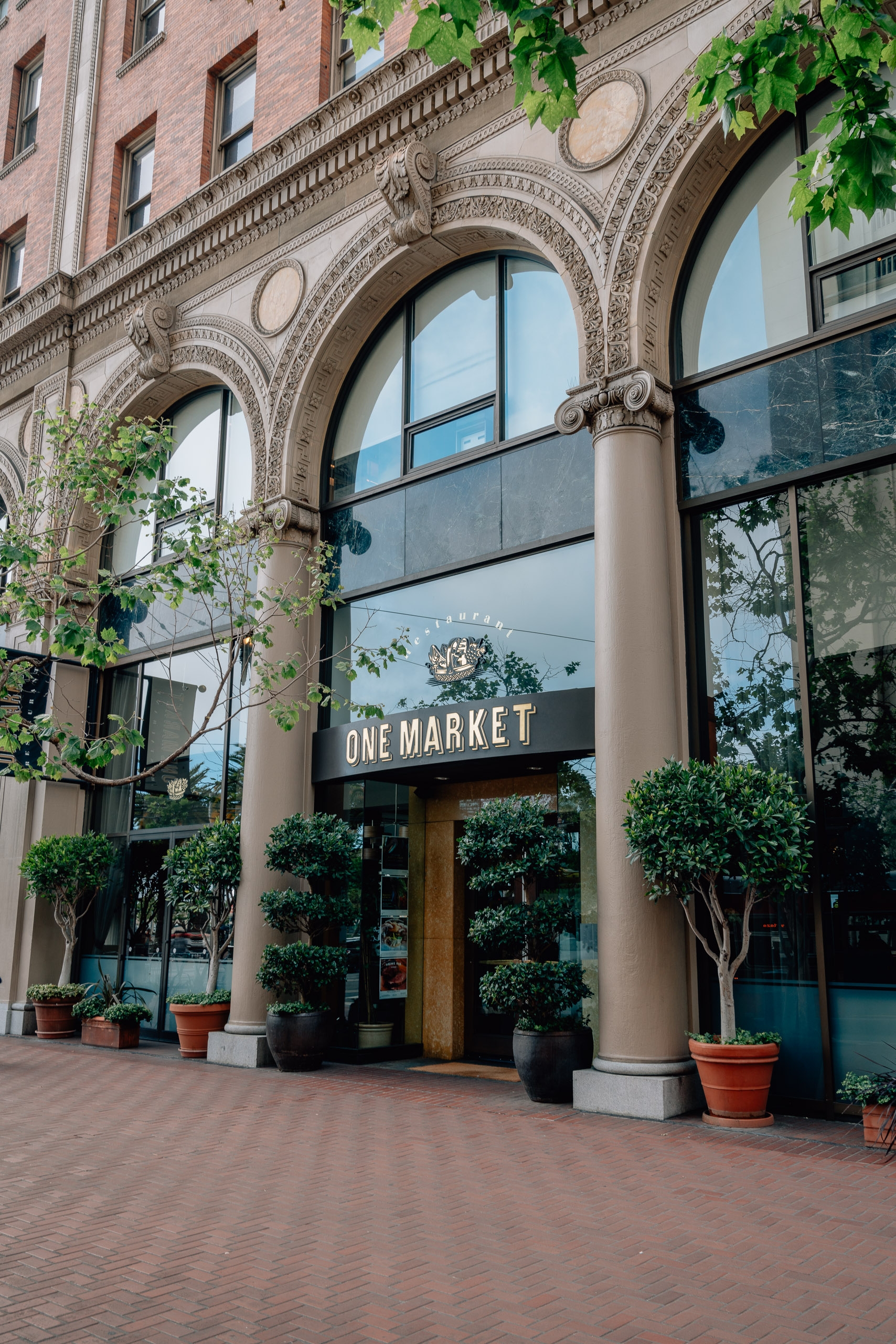 A brick storefront with ornate architectural details houses "One Market," framed by large arched windows and flanked by potted topiaries under afternoon sunlight
