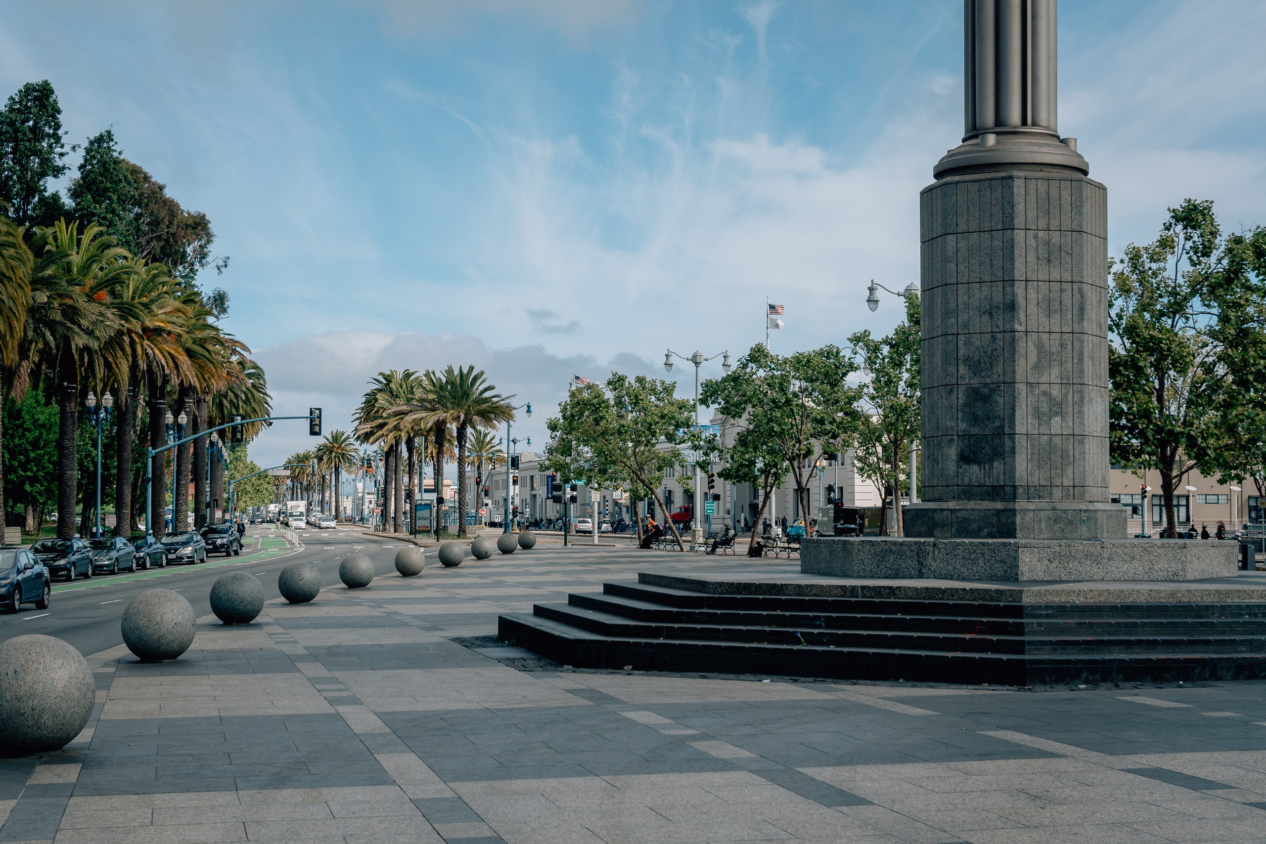 A wide public plaza features a tall cylindrical monument with palm trees lining both sides, spherical bollards in the foreground, and clear blue sky overhead with scattered clouds