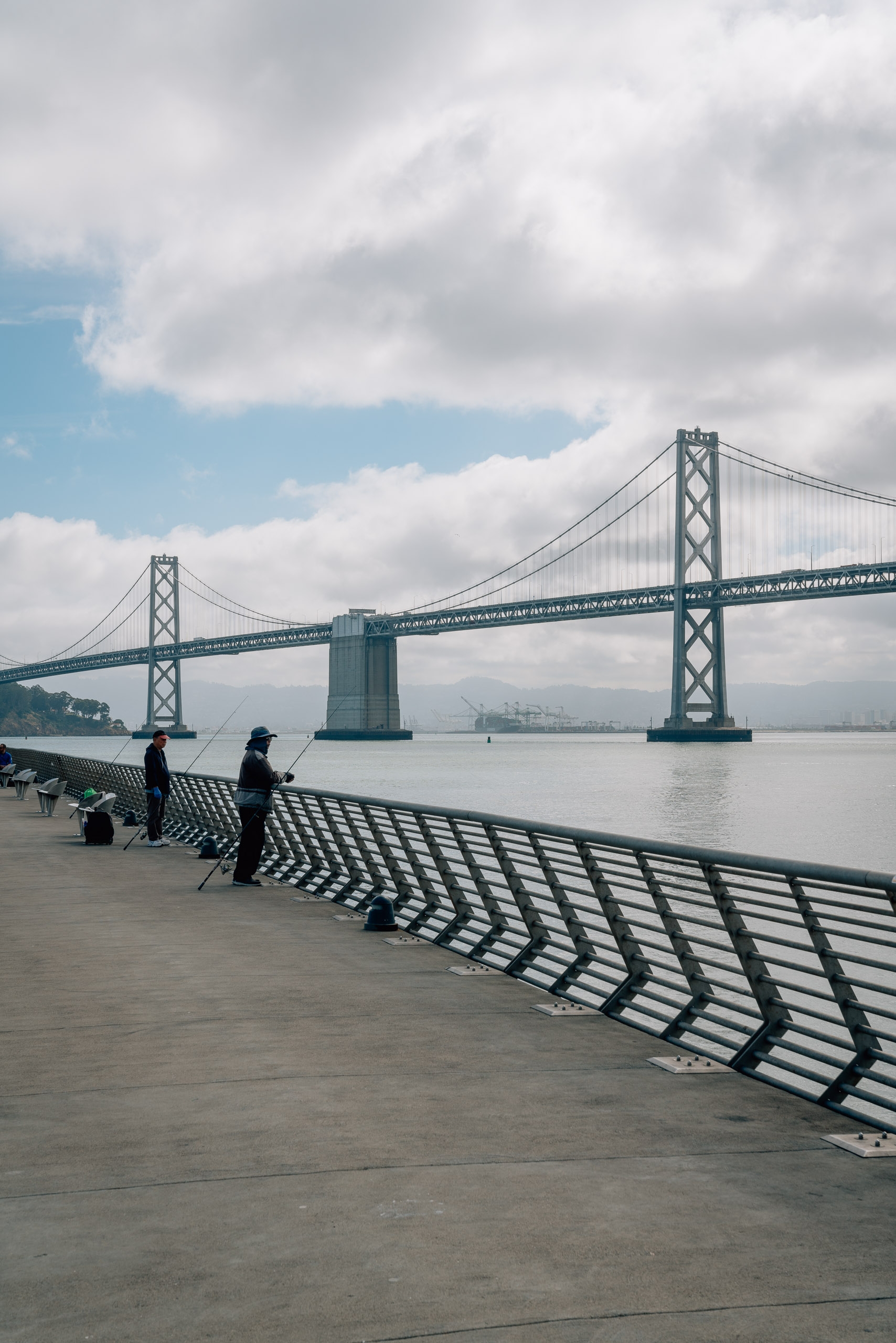People sit on a geometric metal bench along a concrete waterfront promenade facing the Bay Bridge under partly cloudy skies and distant hills