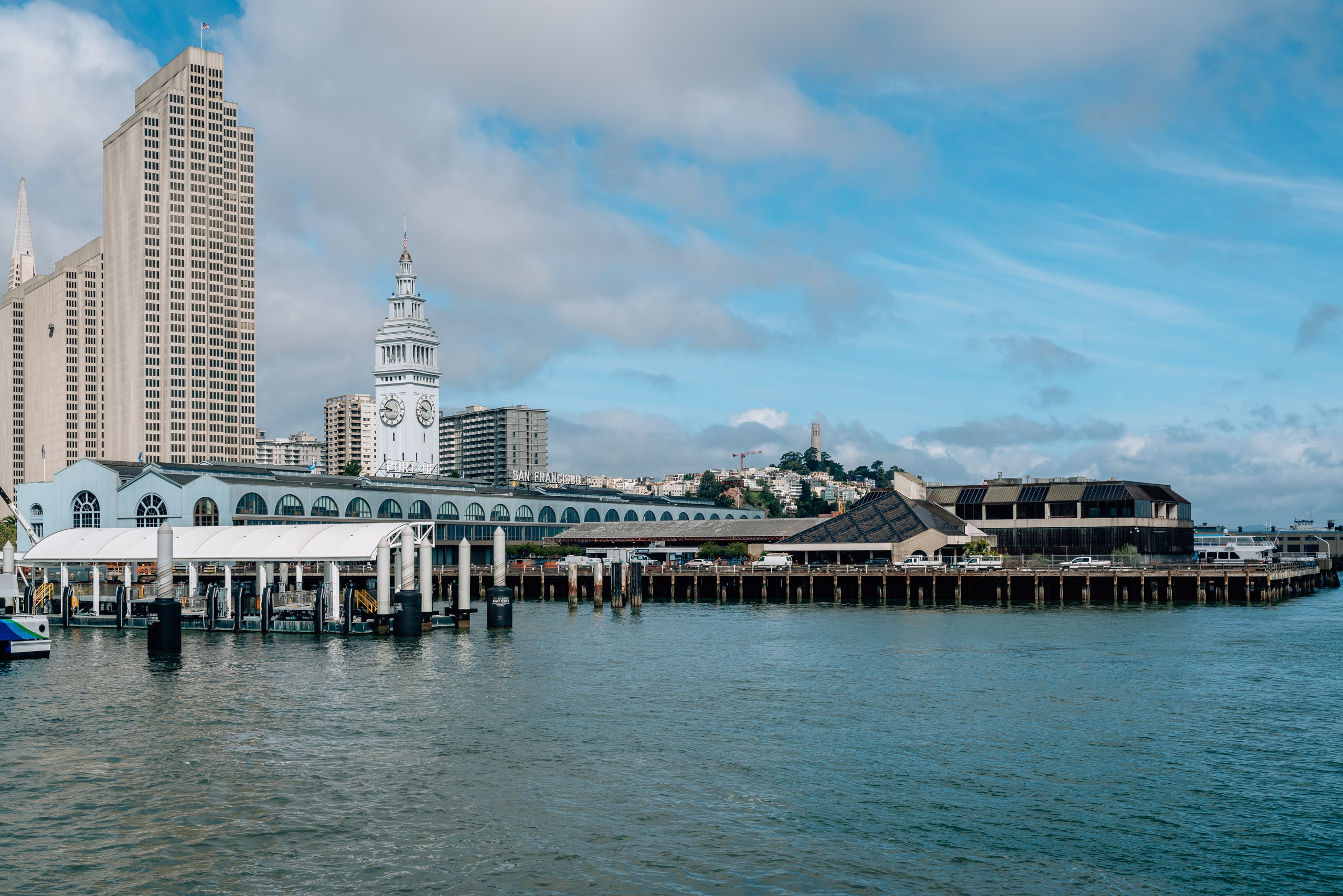 San Francisco's waterfront features the iconic Ferry Building with its clock tower, modern high-rises, and wooden piers extending into turquoise bay waters under partly cloudy skies