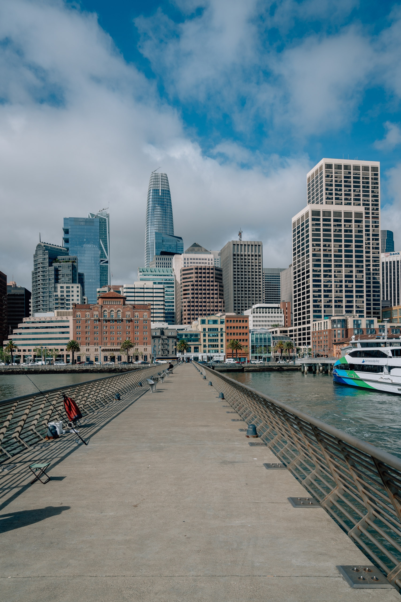 A wooden pier extends toward San Francisco's skyline under bright blue skies, with modern high-rises, the distinctive Transamerica Pyramid, and a moored ferry boat visible across the bay