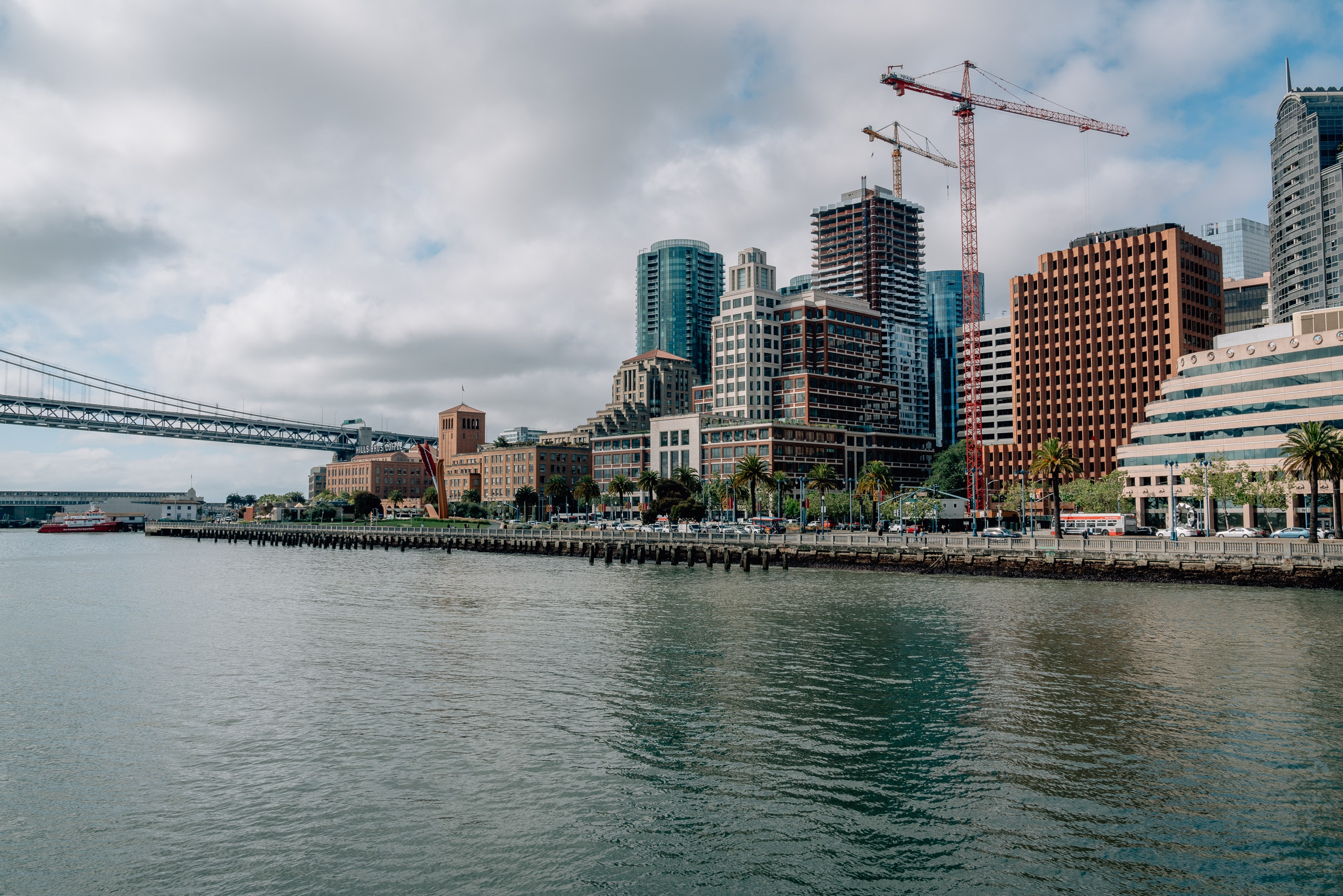 San Francisco's waterfront skyline rises along the bay with construction cranes, mixed architecture, and a steel bridge visible under cloudy skies