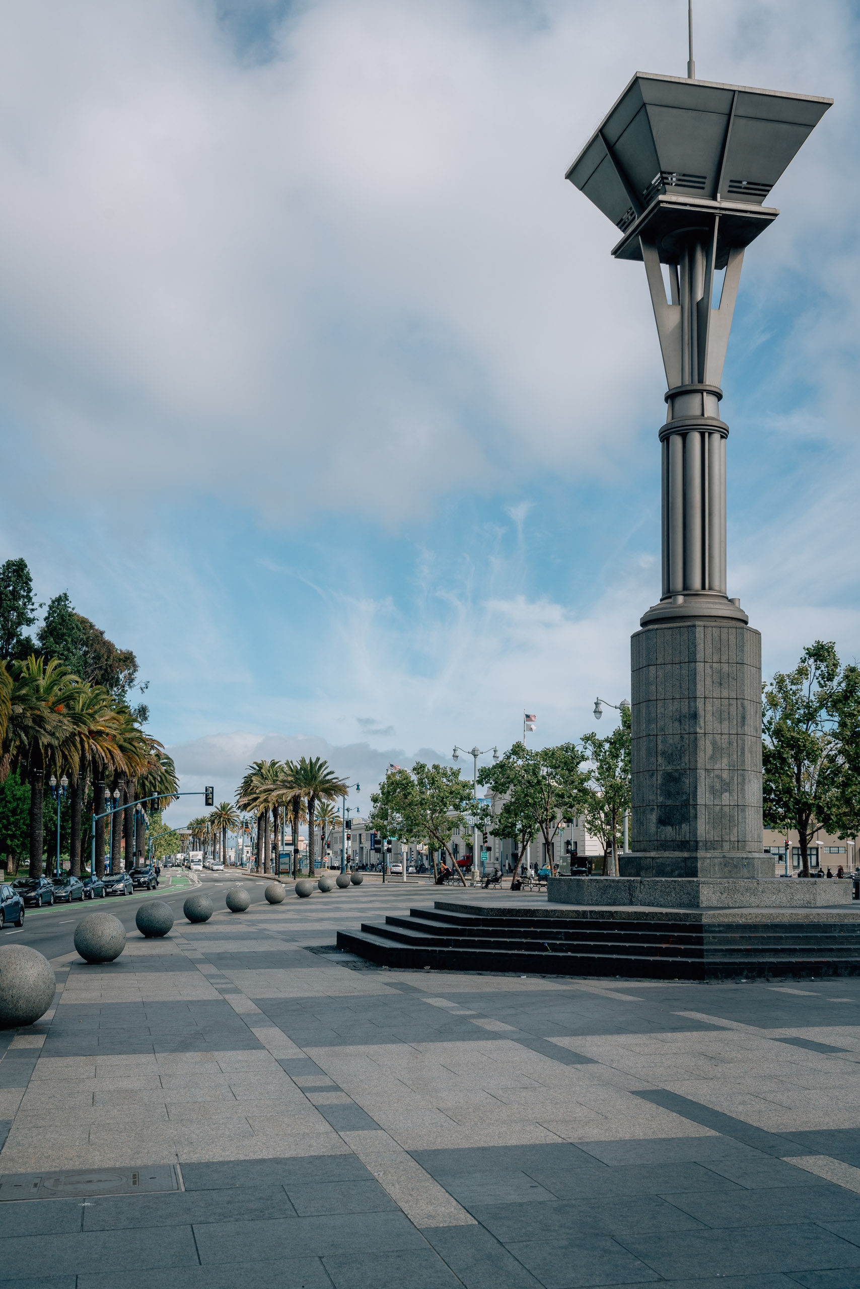 A tall modern street lamp with solar panels stands in a wide plaza lined with palm trees under a partly cloudy sky in San Francisco's Embarcadero district
