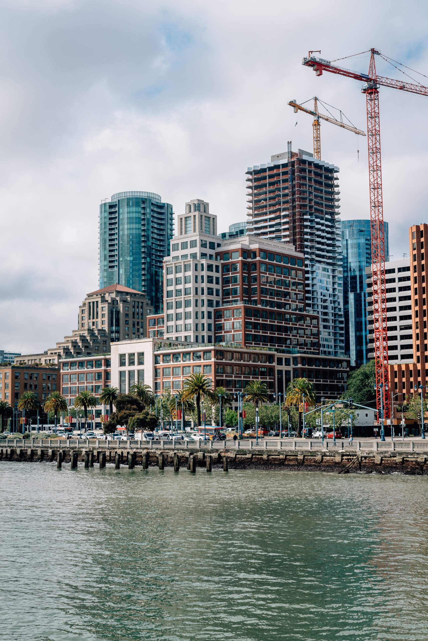 A waterfront cityscape showing mixed architectural styles along a pier with construction cranes overhead and calm green water below under cloudy skies