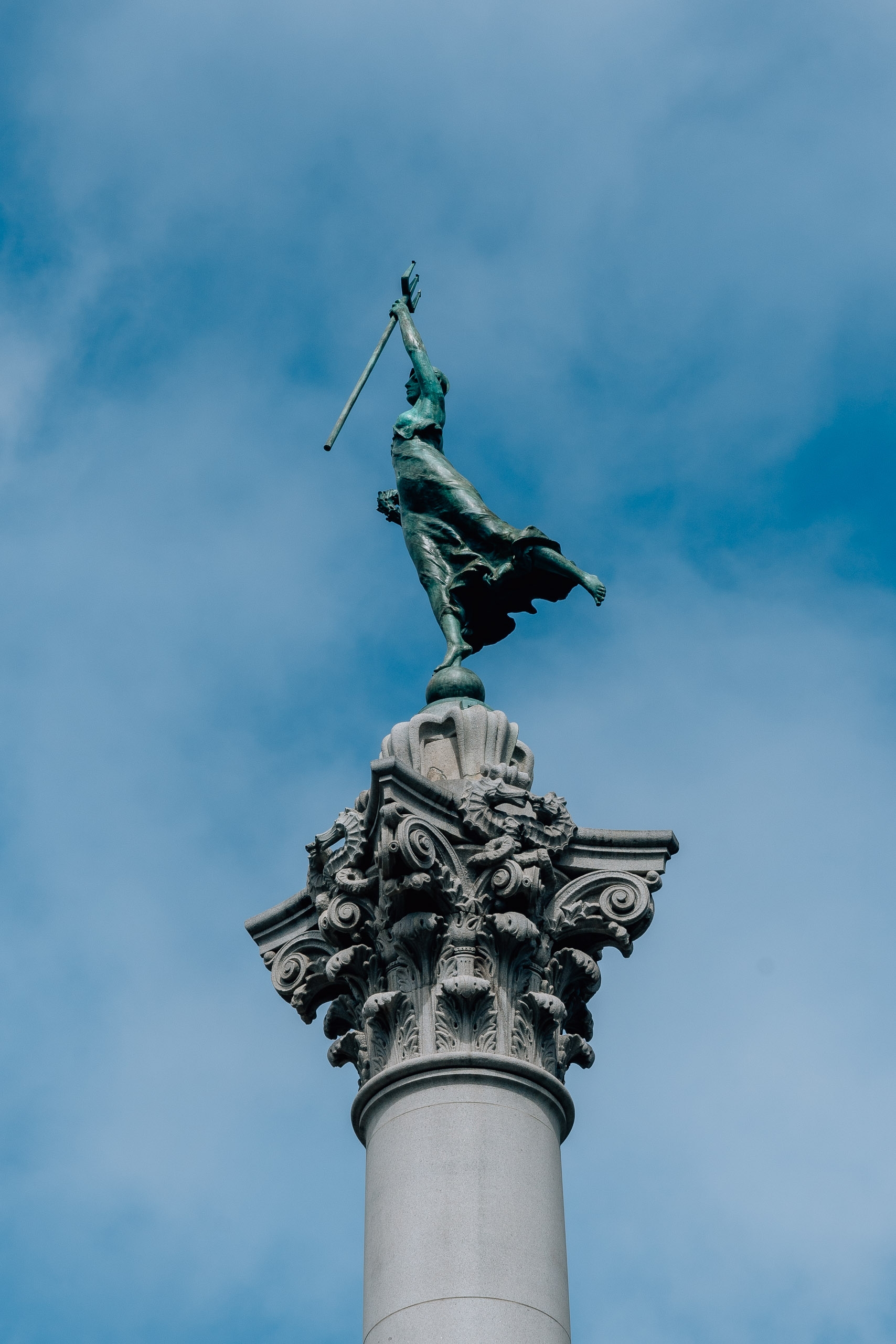 A bronze statue of a winged figure holding a flag stands atop an ornate Corinthian column against a bright blue sky with scattered clouds in San Francisco's Union Square