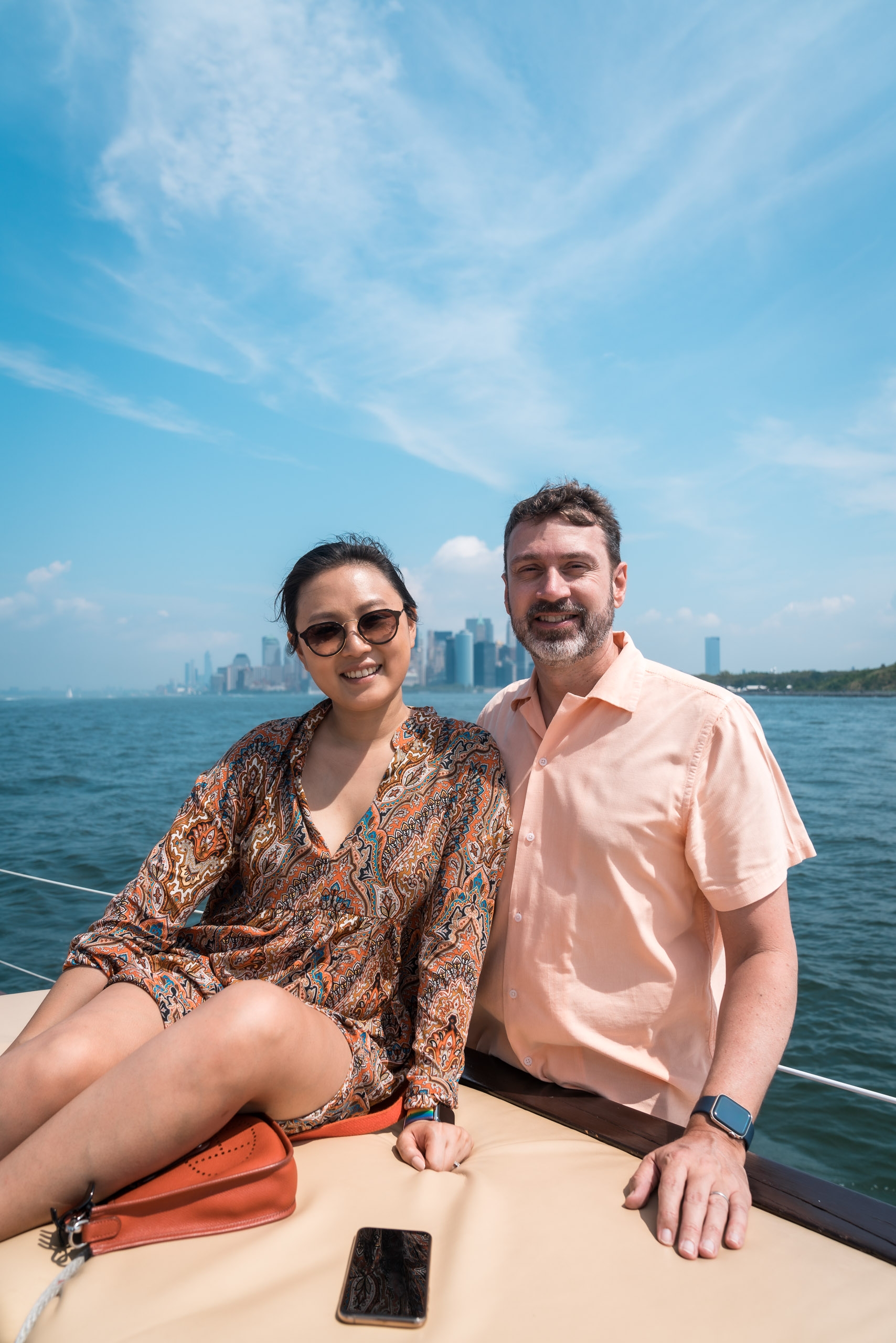 Two people sit on a boat deck with Manhattan's skyline visible across the blue water under a partly cloudy sky