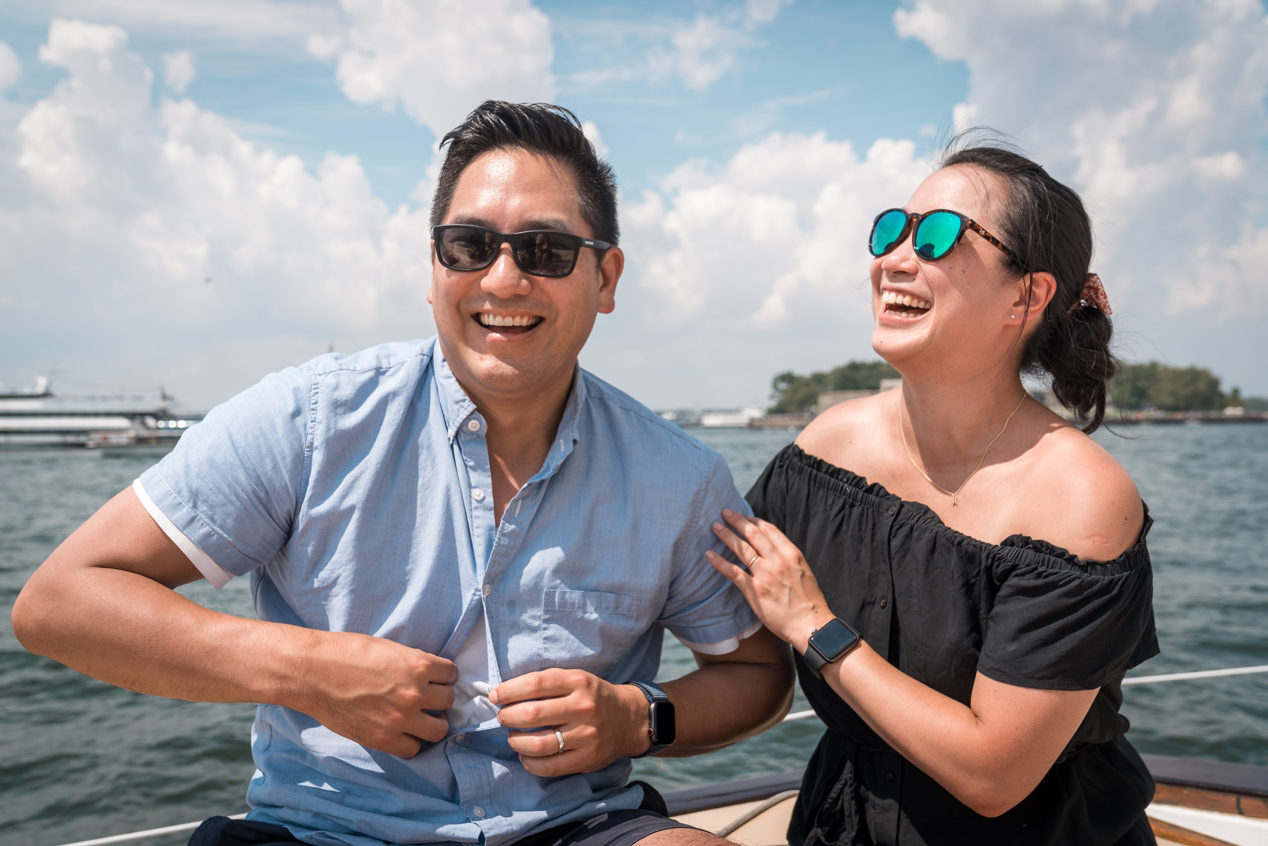 Two people in sunglasses laugh together on a boat with calm blue water and white clouds behind them under bright daylight