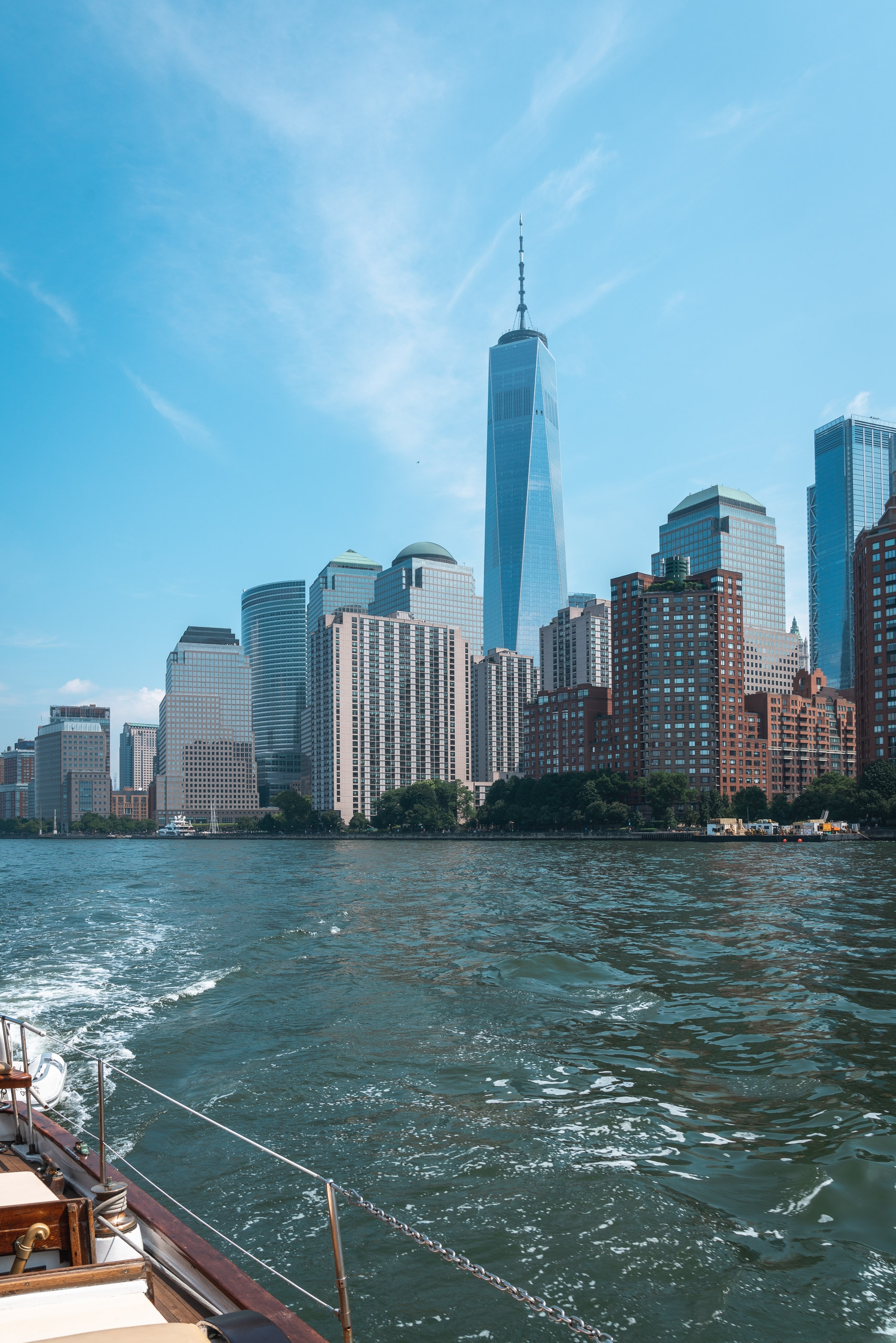 Lower Manhattan skyline rises against clear blue sky, viewed from a boat on the Hudson River with One World Trade Center prominent in the center