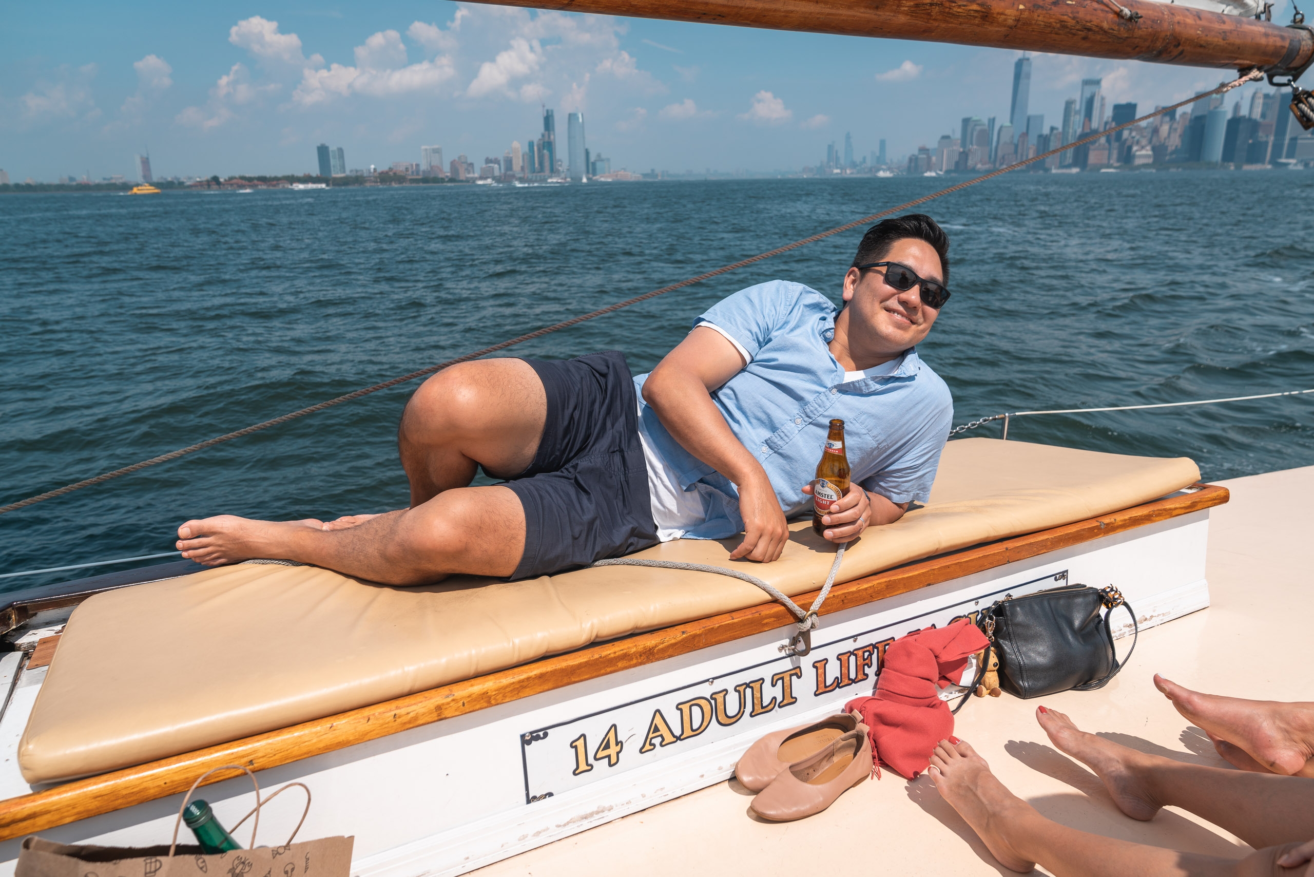 A man relaxes on a sailboat's cushioned bench with a beer bottle while Manhattan's skyline glimmers across the water under bright daylight