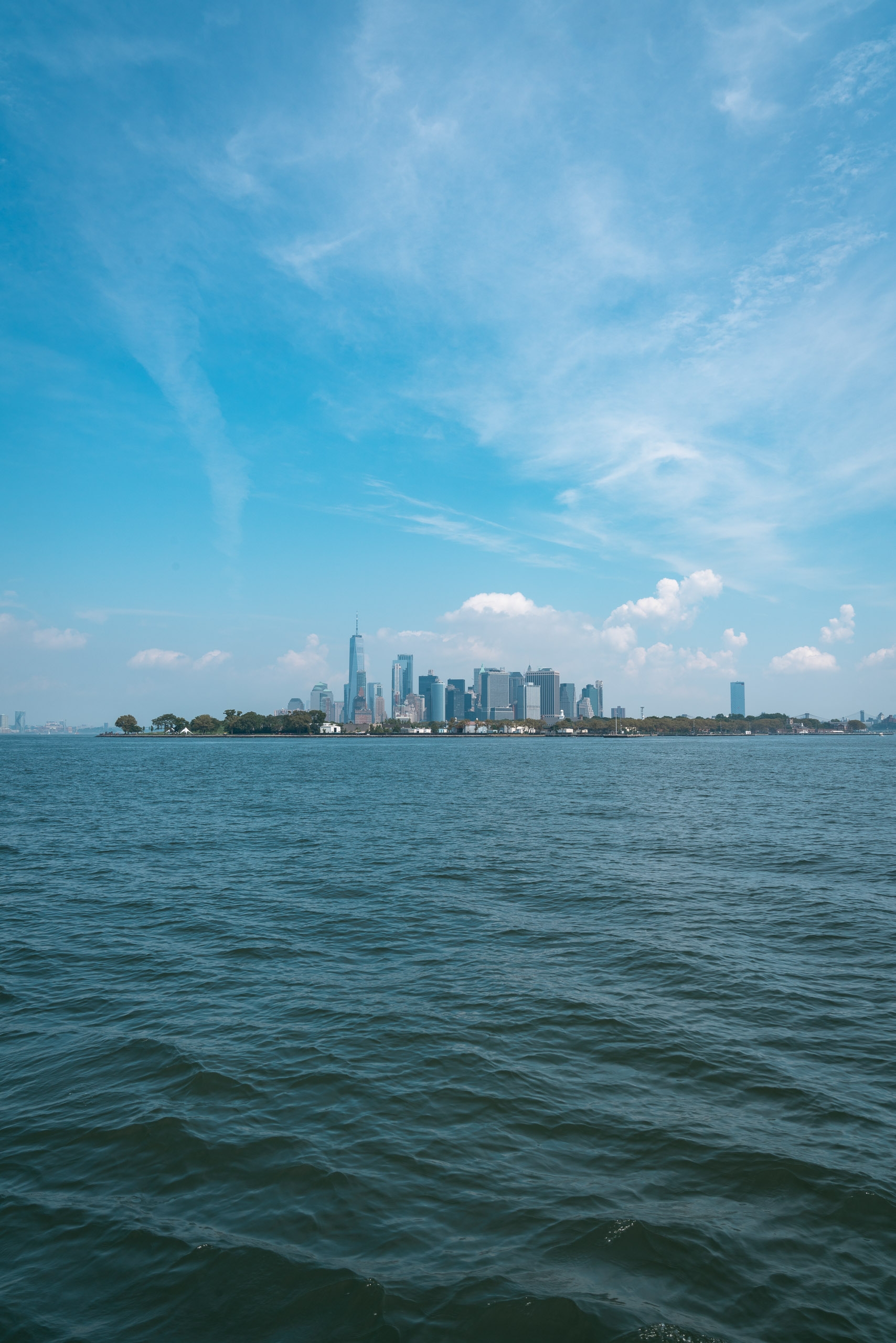 A distant Manhattan skyline rises above calm green water under a bright blue sky dotted with white clouds