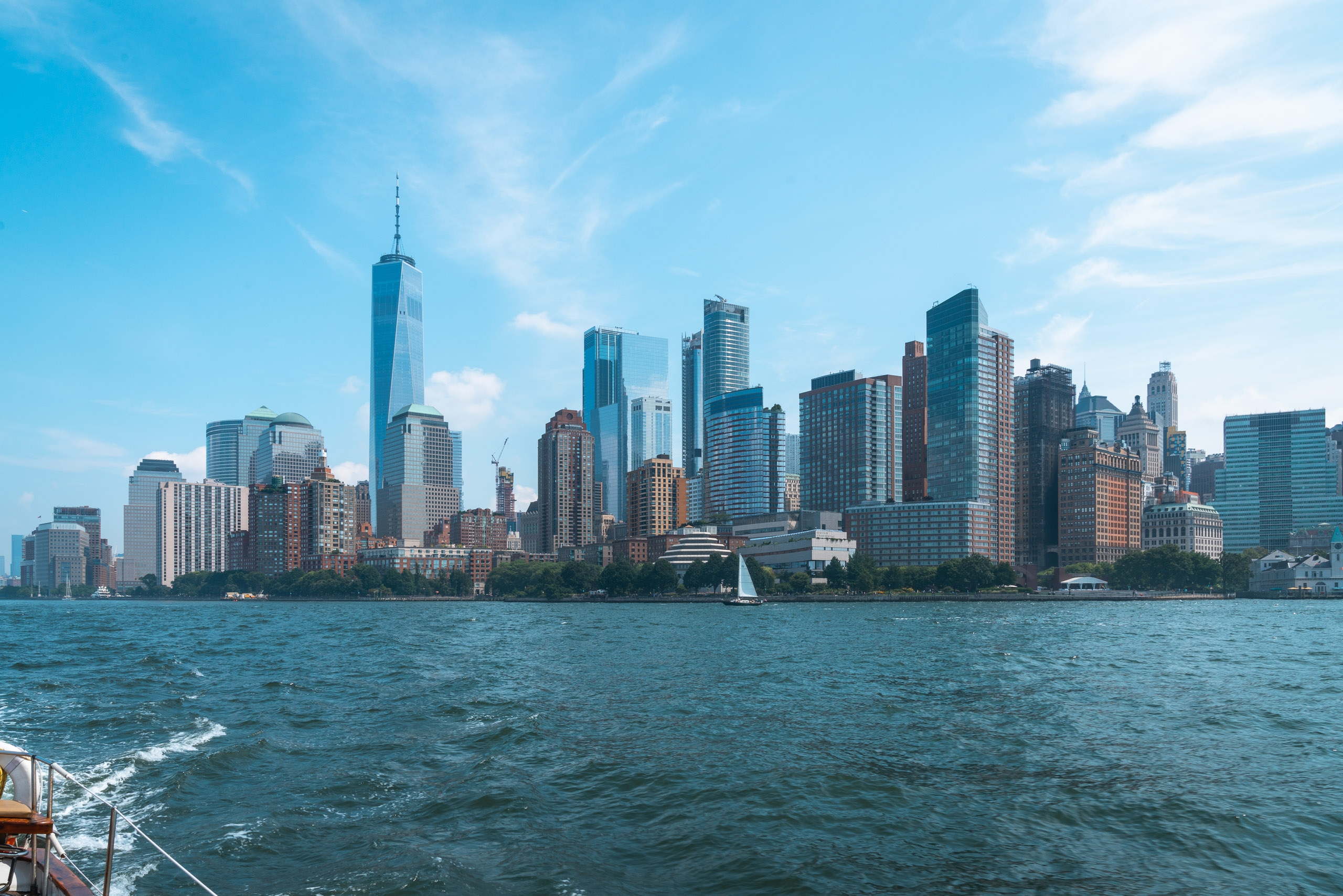 Manhattan skyline viewed from the water on a clear day, with One World Trade Center rising prominently among diverse skyscrapers under bright blue sky and white clouds