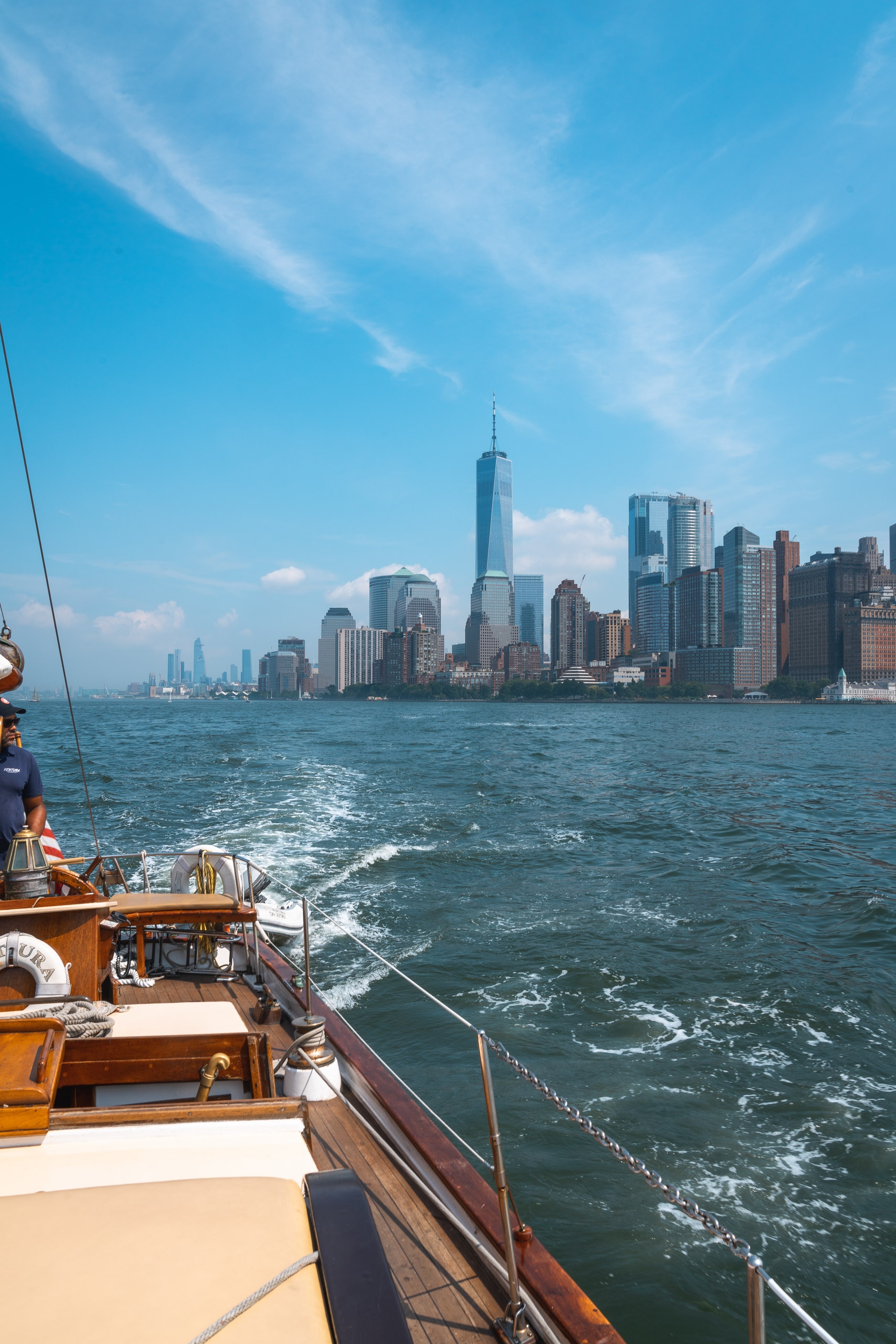 A wooden sailboat approaches Manhattan's skyline across choppy harbor waters under clear blue sky, with One World Trade Center prominent among the city's towering buildings