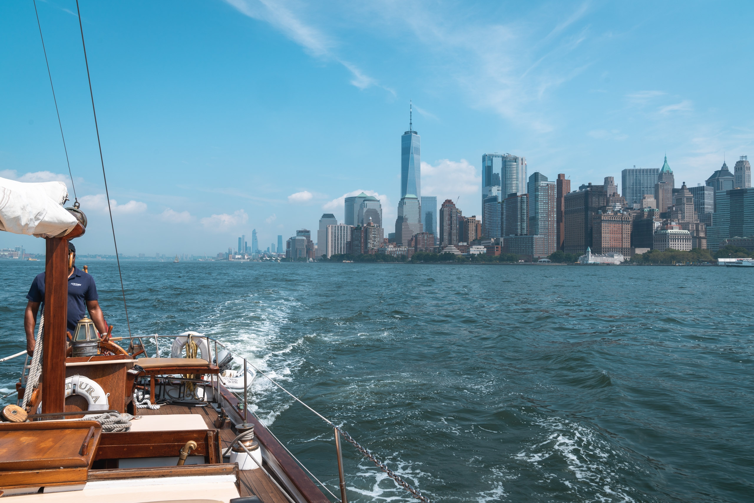 A wooden sailboat approaches Manhattan's skyline across calm blue-green water under clear sky, with the boat's mast and rigging visible in the foreground