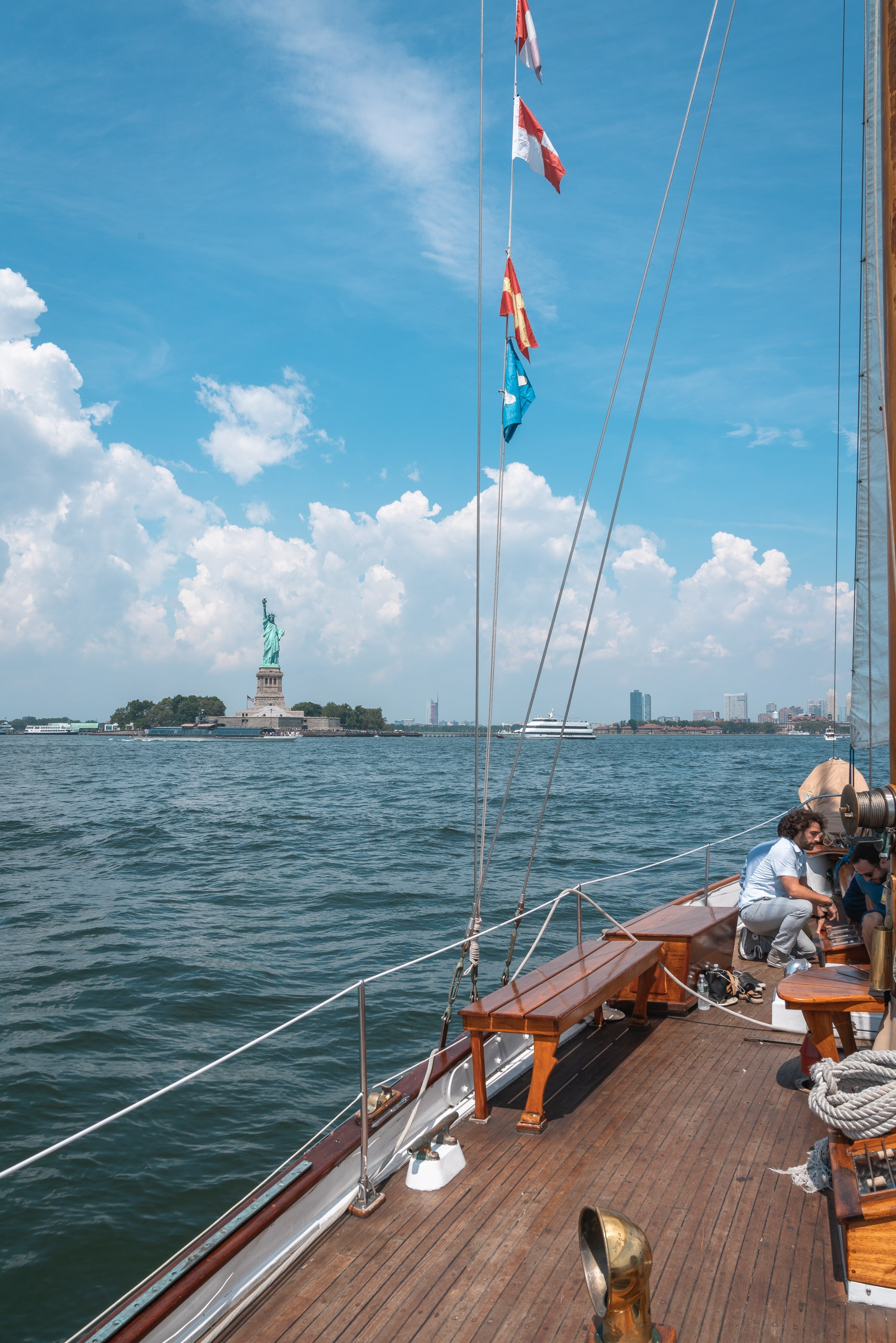A sailboat's wooden deck stretches toward the Statue of Liberty under blue sky with white clouds, flags fluttering from the mast above calm harbor waters