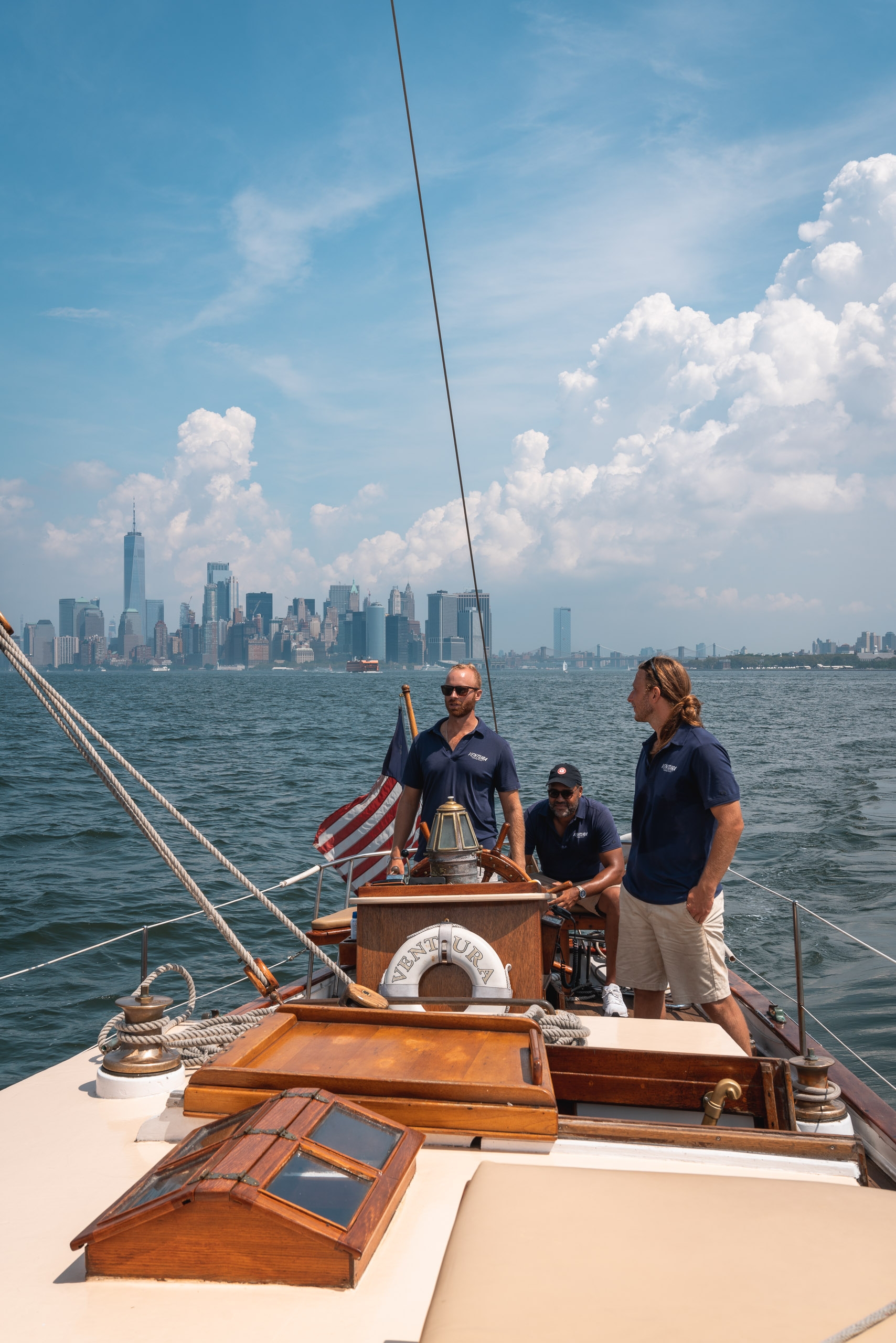 Three crew members stand on a wooden sailboat's deck with Manhattan's skyline visible across the water under a partly cloudy blue sky