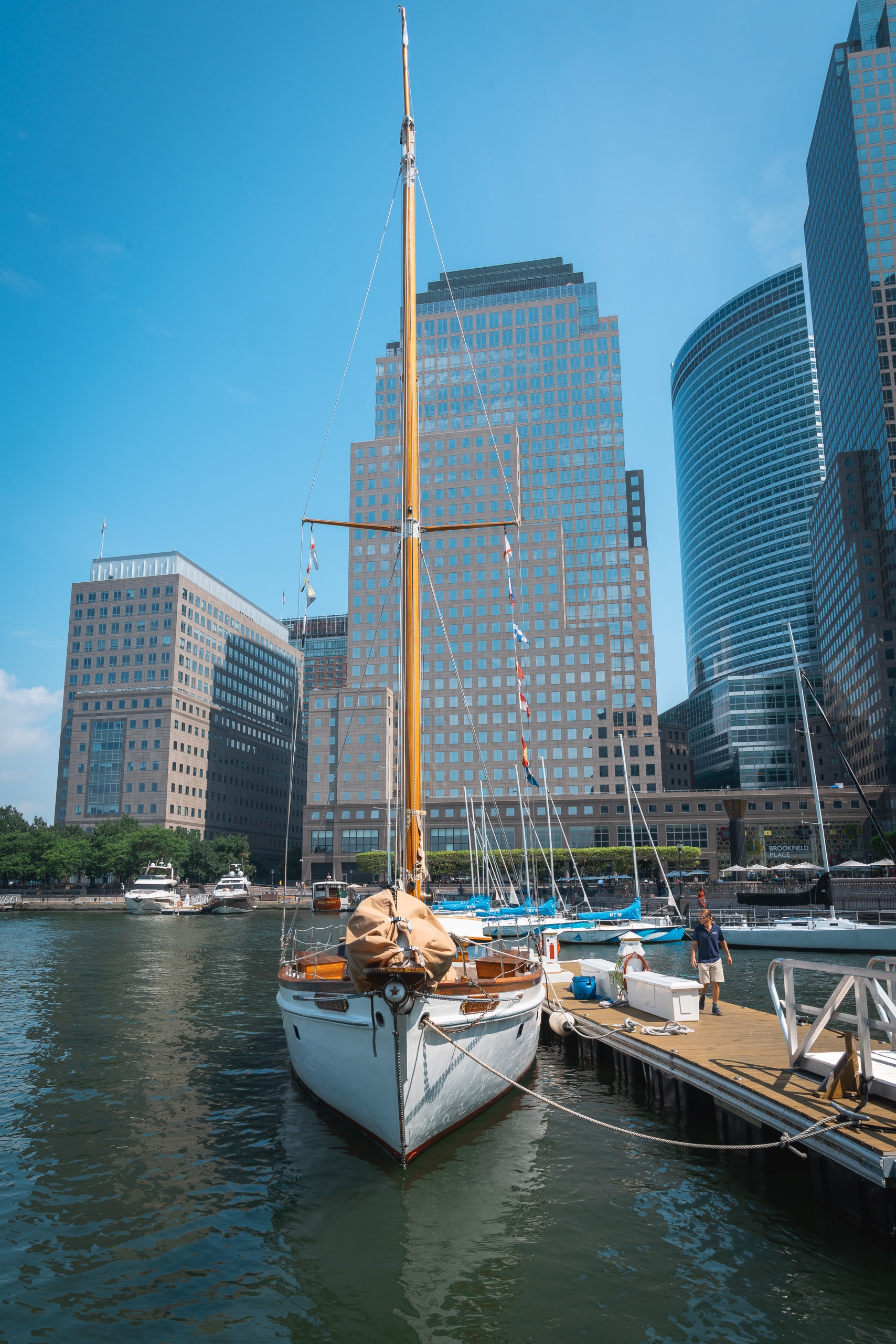 Wooden sailboat with tall mast docked at a Manhattan waterfront pier, modern glass skyscrapers rising behind calm water under clear blue sky