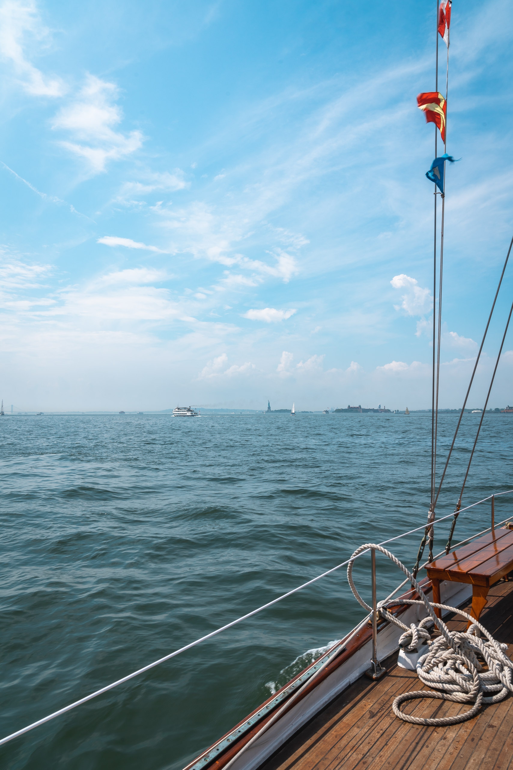 A wooden sailboat's deck extends toward calm waters with red and yellow flags flying from the mast, distant Manhattan skyline visible across the bay under a bright blue sky