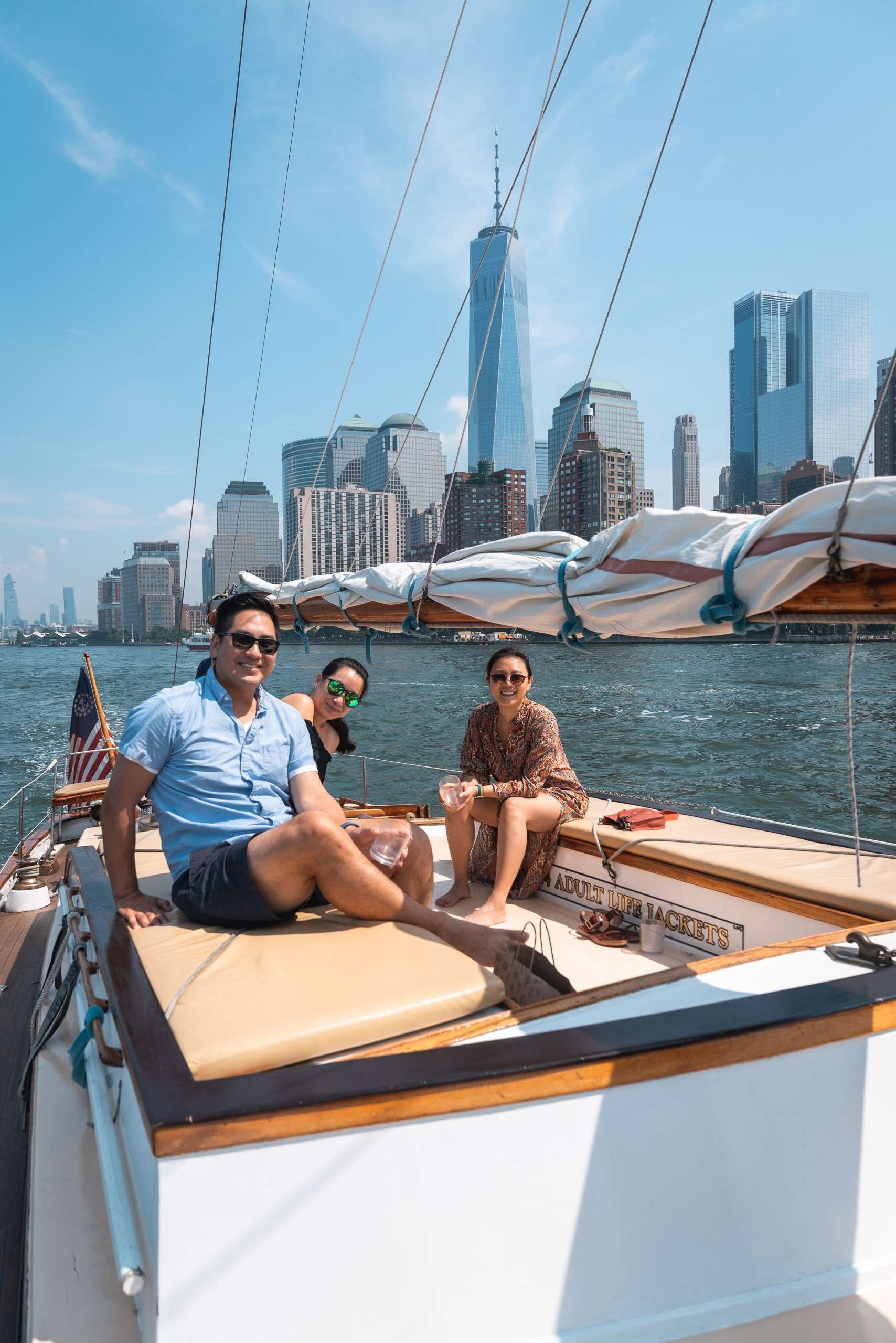 Three people relax on a wooden sailboat with the Manhattan skyline, including One World Trade Center, rising behind them across the blue harbor water