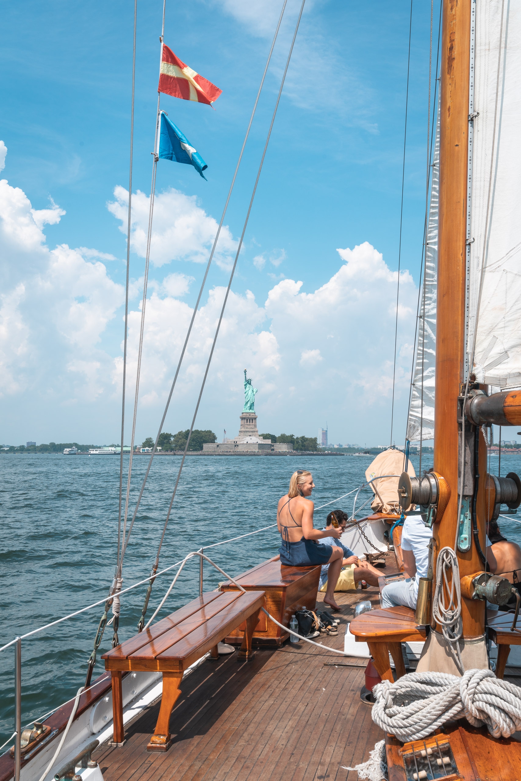 Passengers aboard a wooden sailing vessel navigate past the Statue of Liberty under clear skies, with nautical flags and rigging visible against puffy white clouds