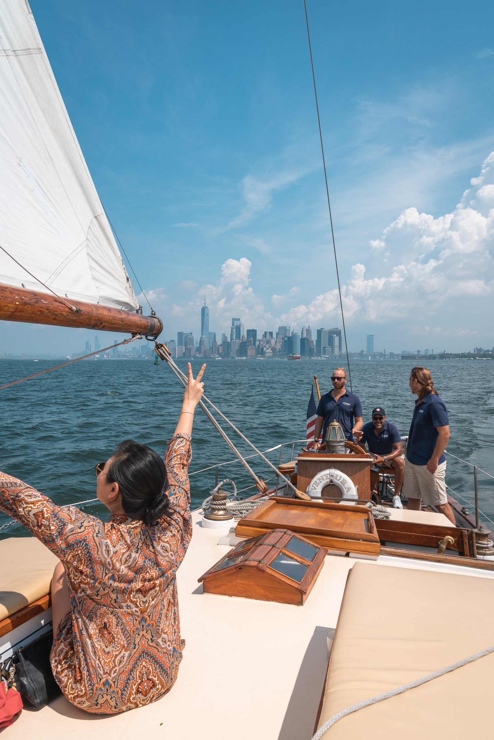 A sailor adjusts rigging on a wooden sailboat with Manhattan's skyline visible across the water under bright blue skies and white clouds