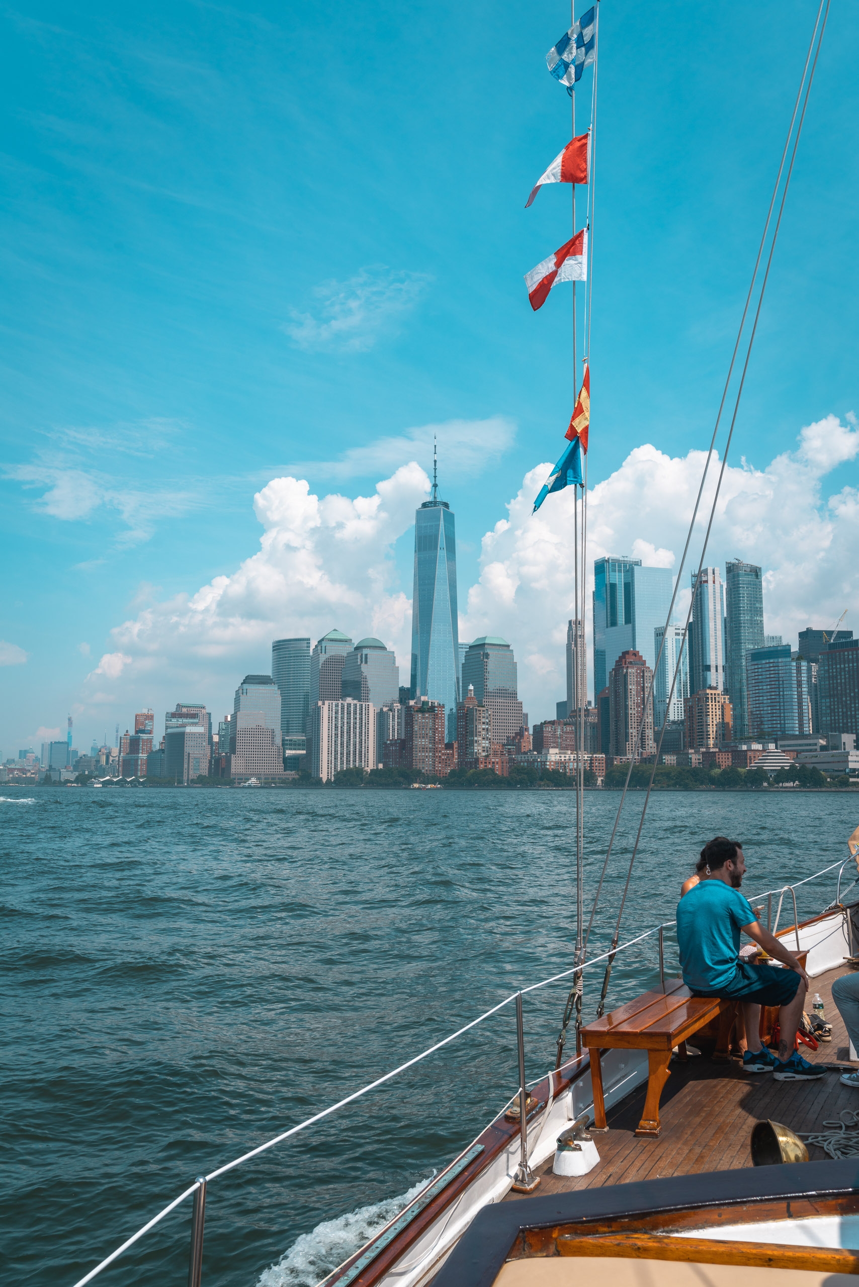 A sailor in a turquoise shirt sits on the wooden deck of a sailboat with international flags flying, gazing toward the Manhattan skyline across turquoise-blue water under a bright sky