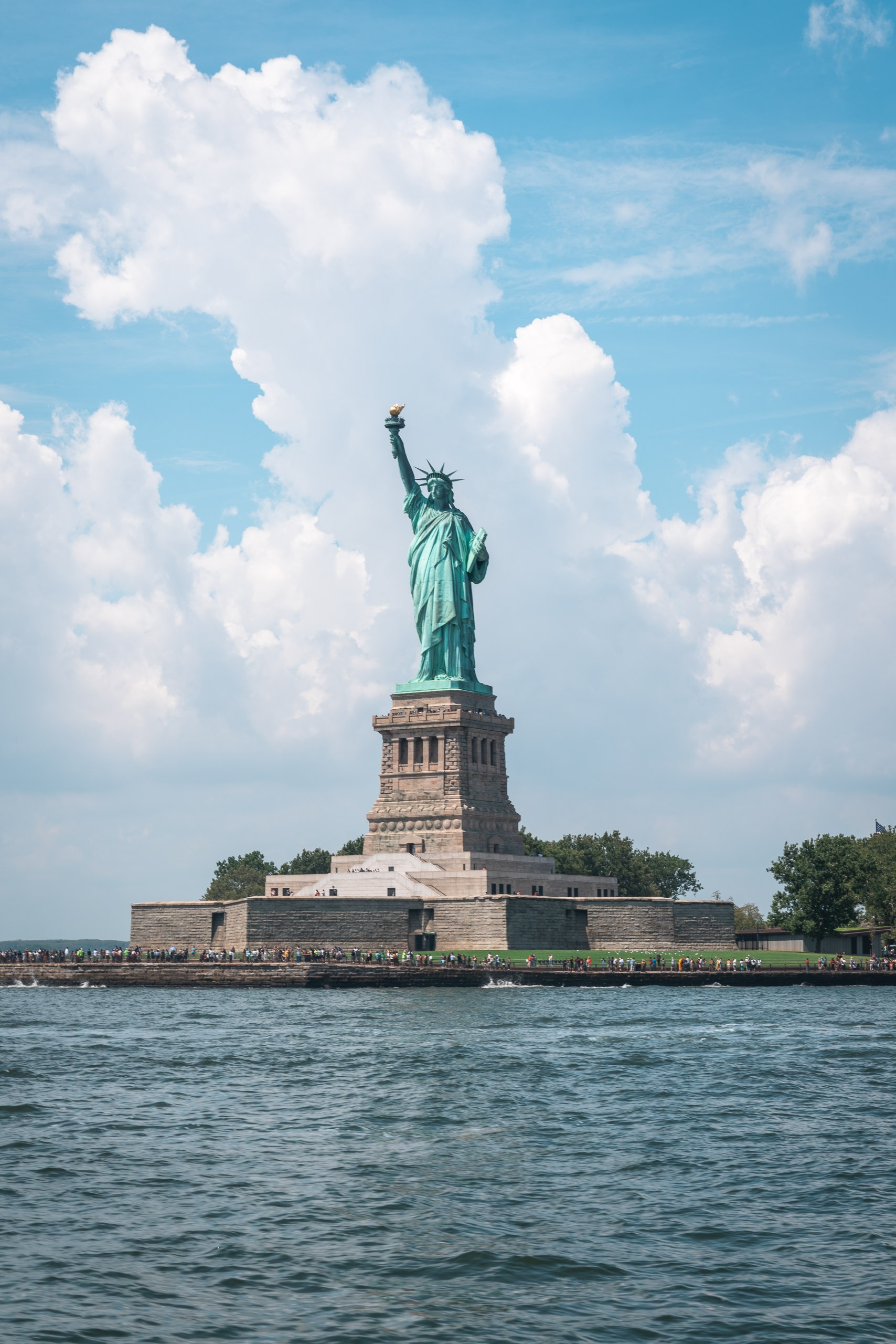 The Statue of Liberty stands on its stone pedestal on Liberty Island, surrounded by water under a bright blue sky with white puffy clouds