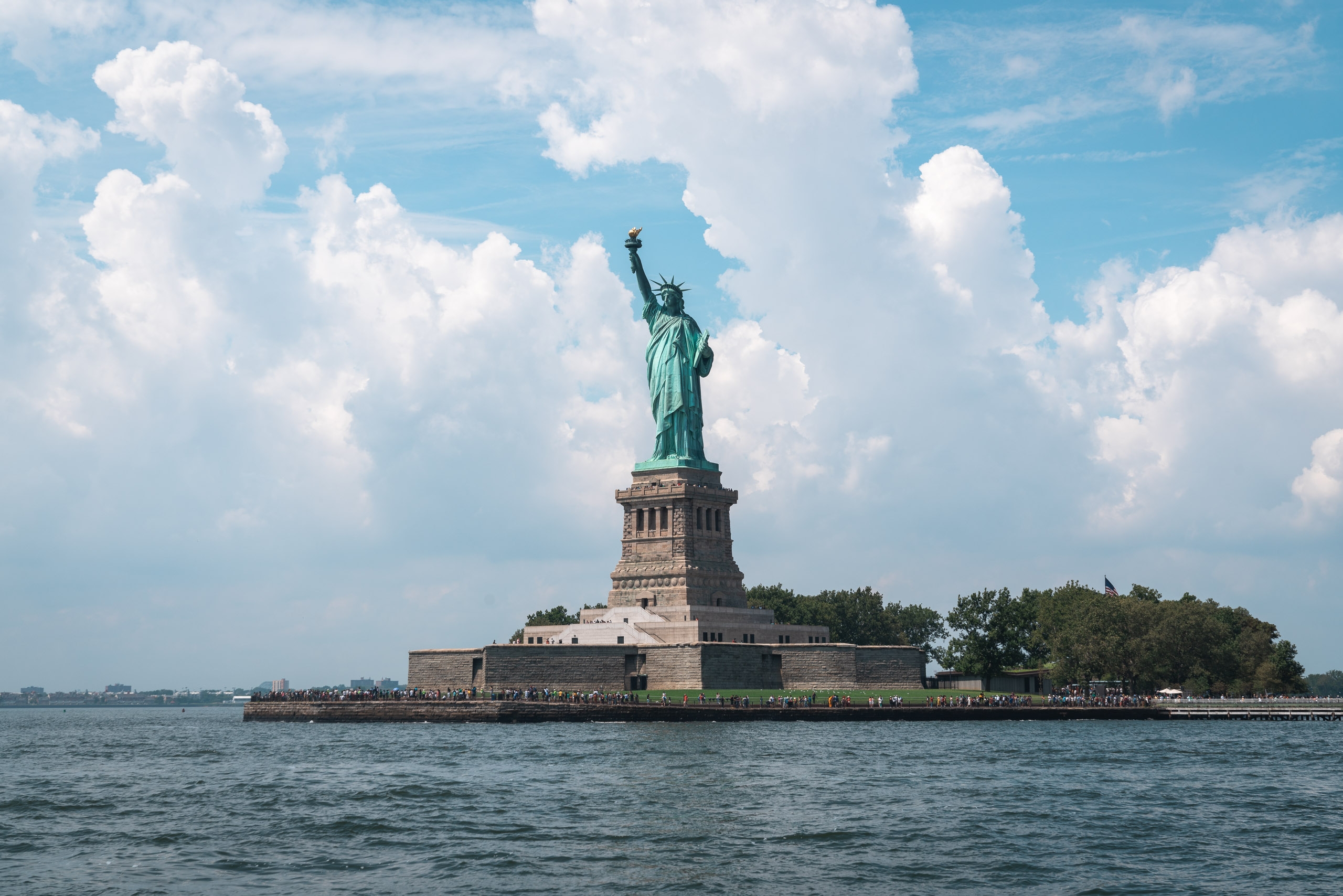The Statue of Liberty stands on its pedestal on Liberty Island, surrounded by water under a bright blue sky dotted with white cumulus clouds