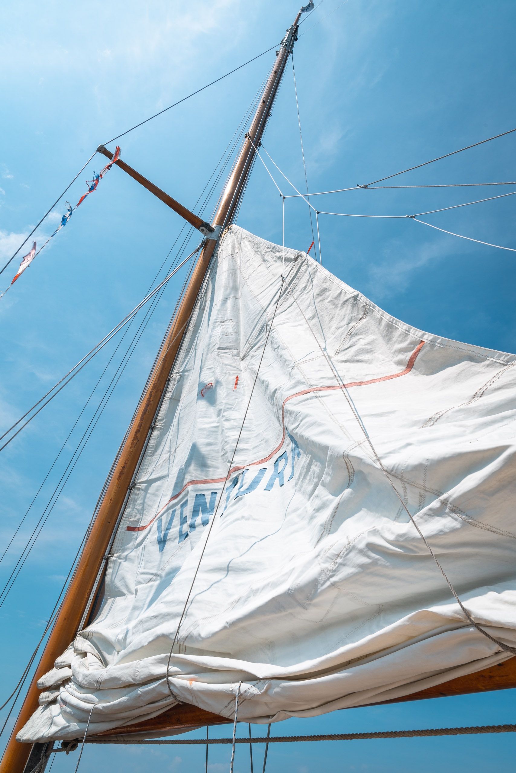 Billowing white sail with blue text catches bright sunlight against a clear sky, wooden mast and rigging visible from below on a sailing vessel