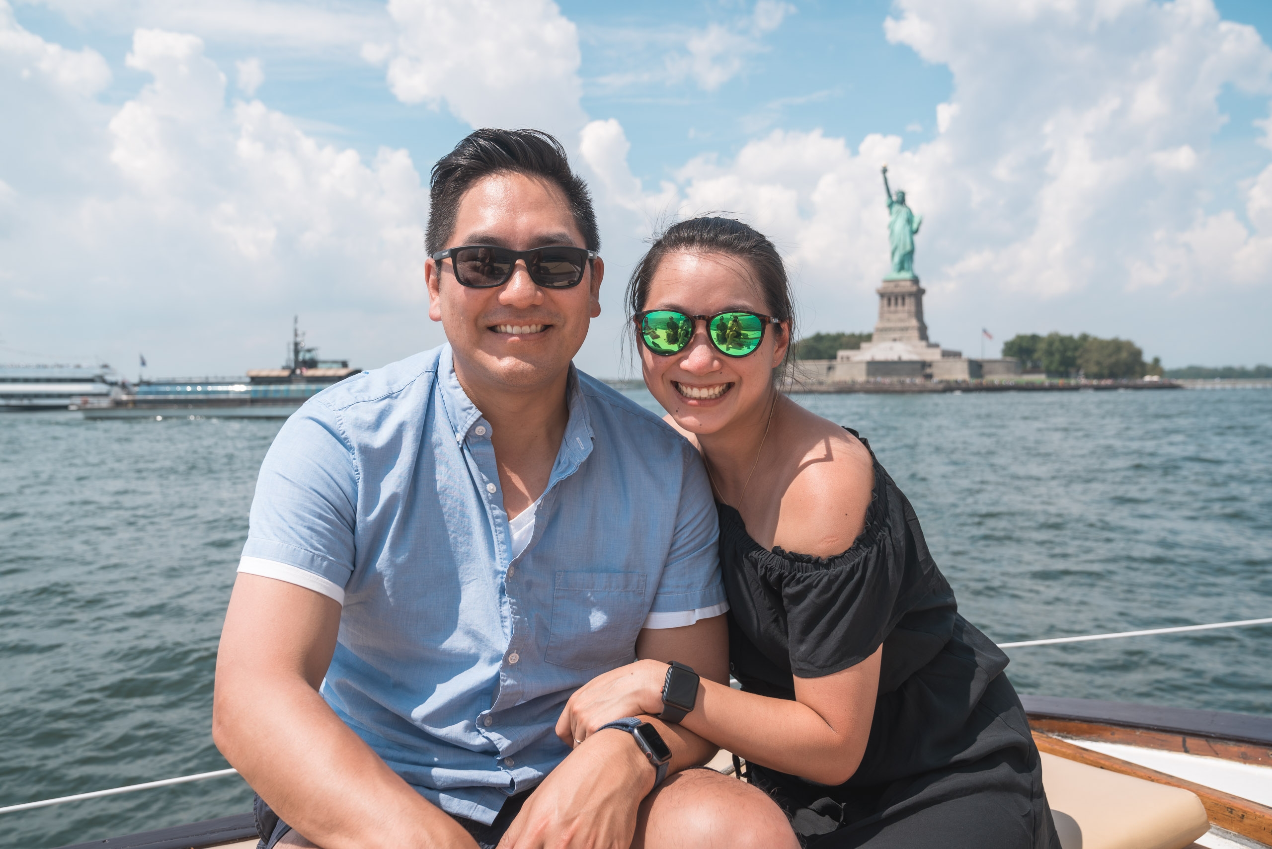 Two smiling visitors pose together on a boat with the Statue of Liberty and Manhattan skyline visible across the water under a bright, partly cloudy sky