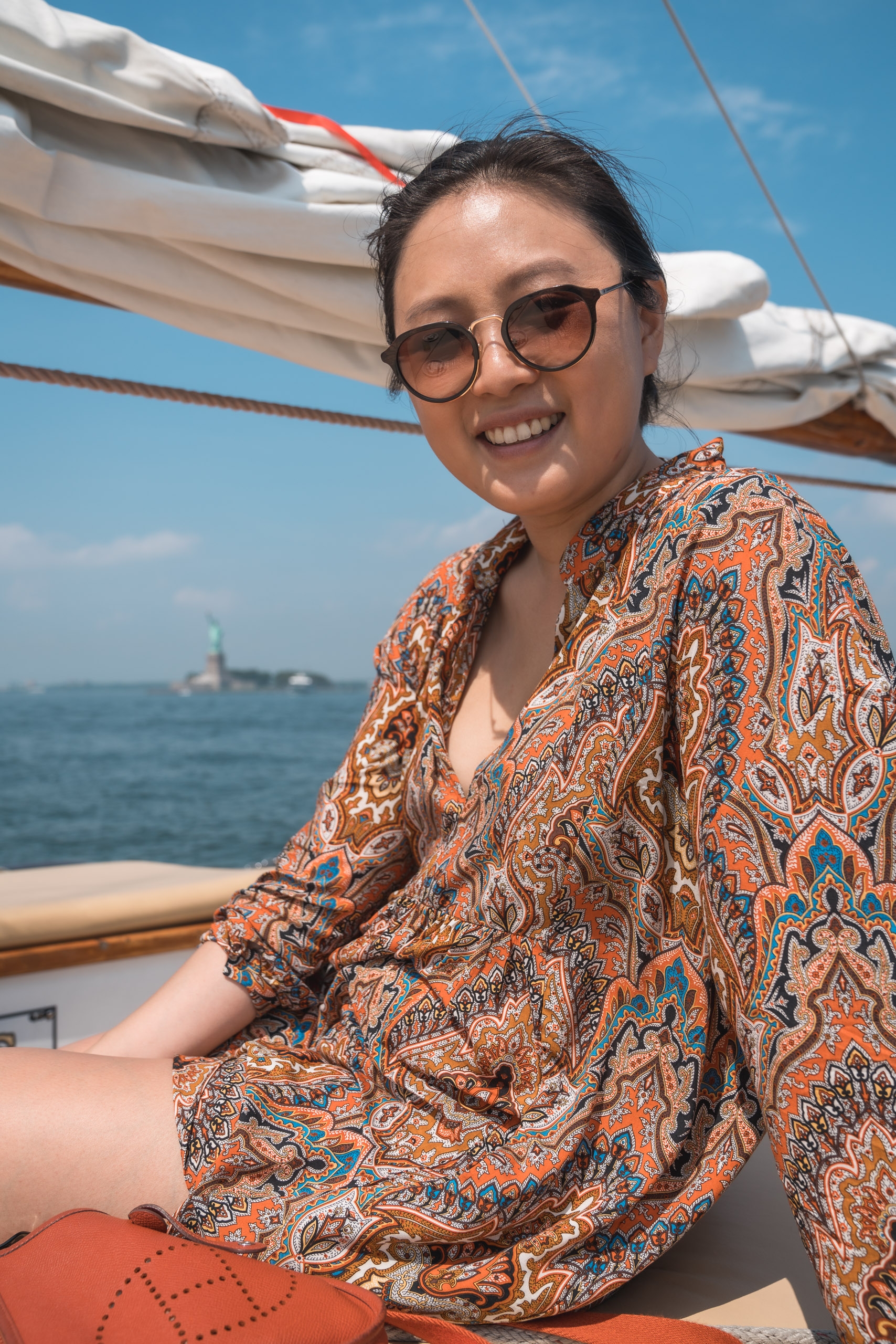A smiling woman wearing sunglasses and an ornate patterned shirt sits on a sailboat deck under blue skies, with white sails and ocean visible behind her