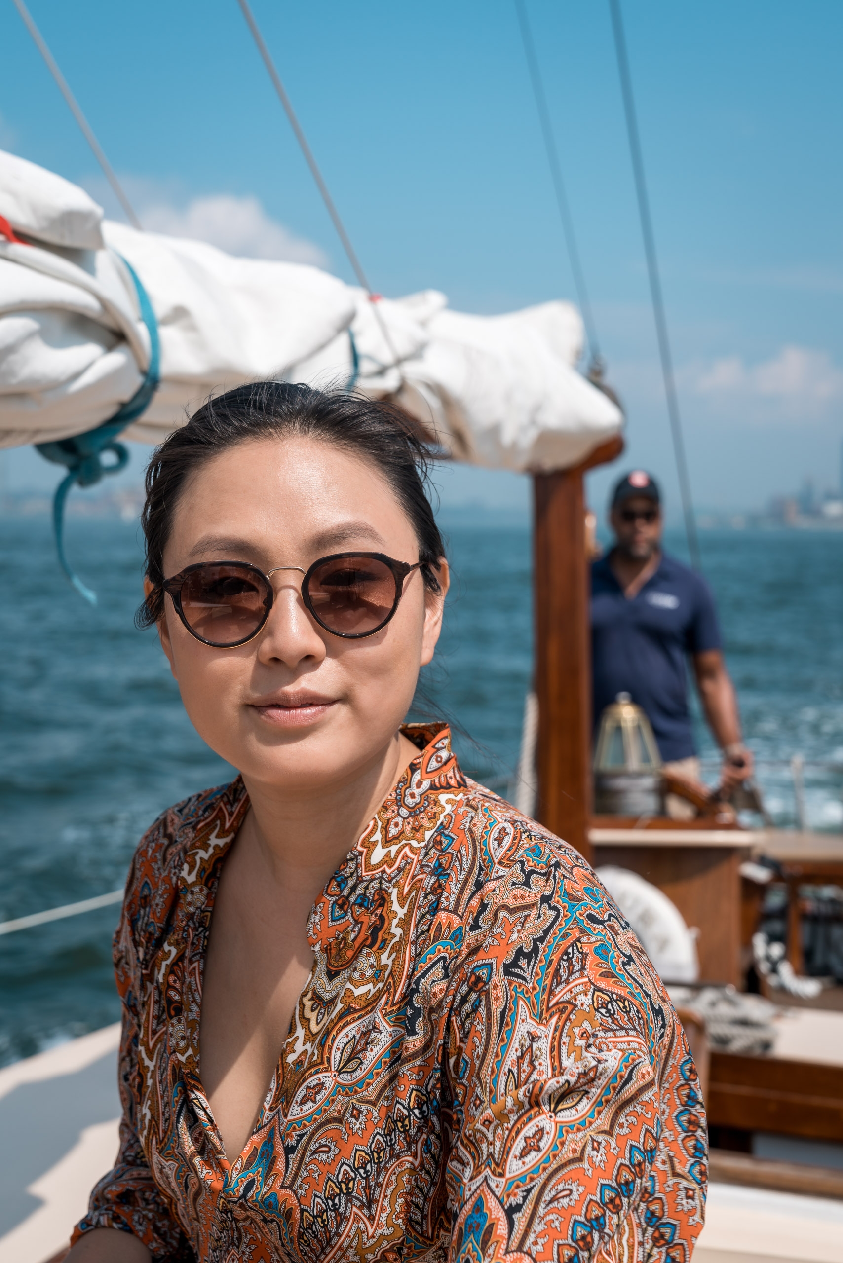 Woman wearing sunglasses and patterned orange shirt poses on a sailboat with white canvas and rigging visible under clear blue sky