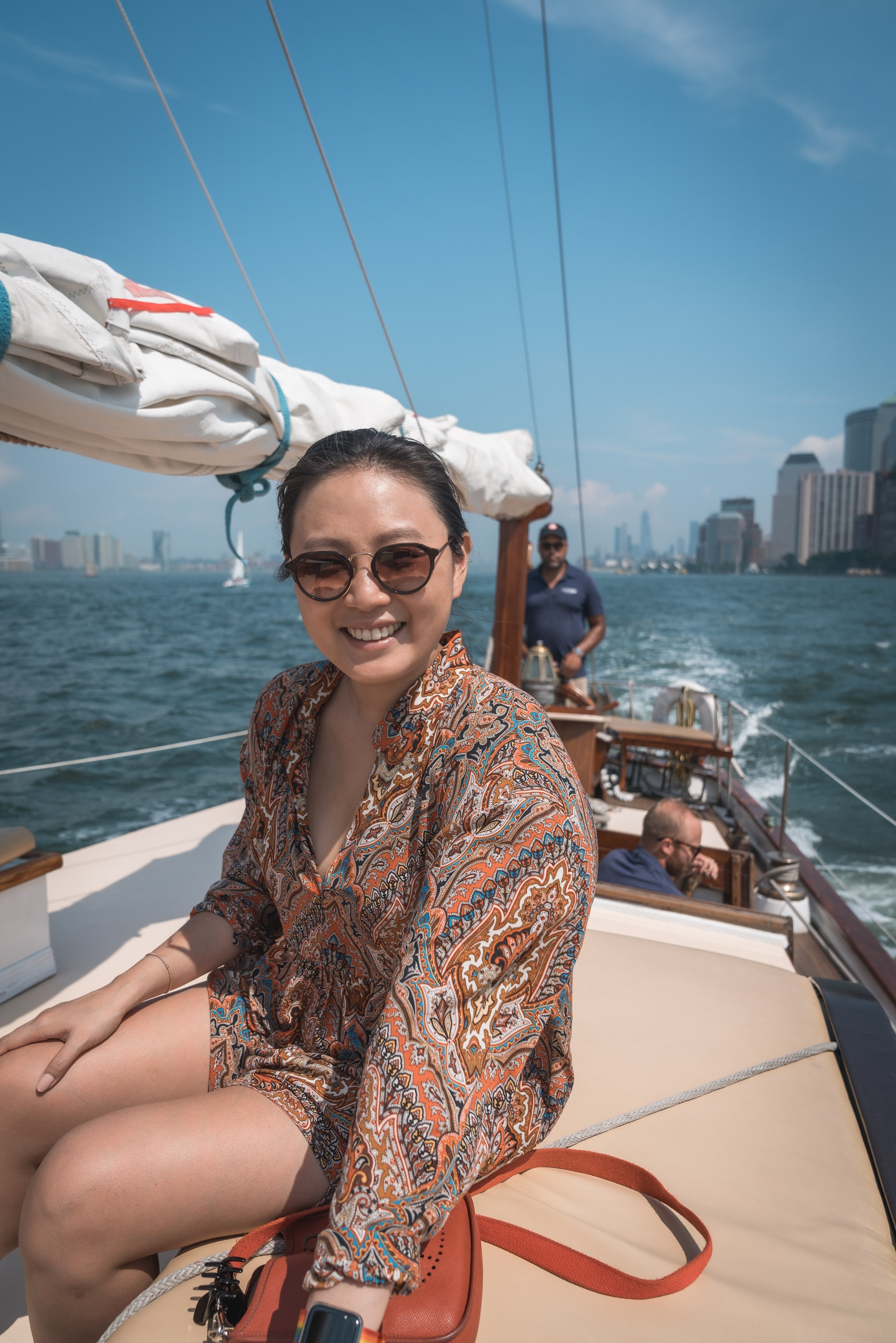 A woman in sunglasses and a patterned shirt smiles at the camera while seated on a sailboat deck with Manhattan's skyline visible across the water behind her