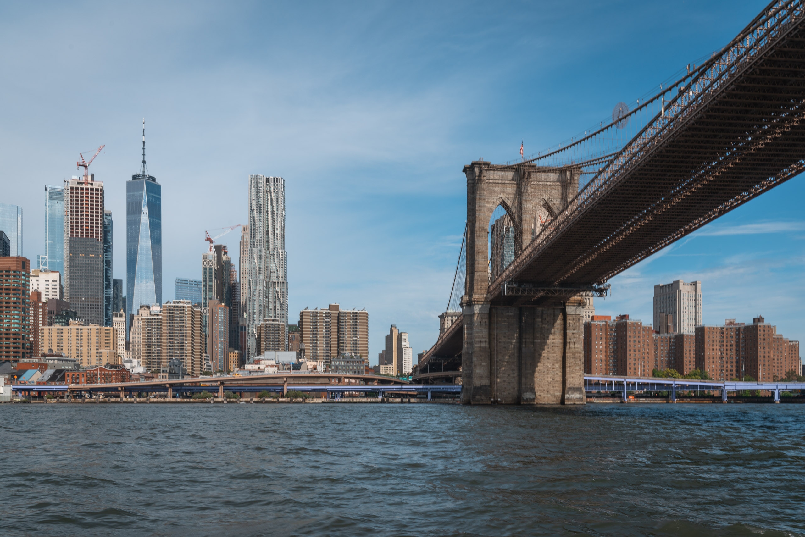 The Brooklyn Bridge spans diagonally across the frame above choppy water, with Manhattan's skyline featuring One World Trade Center and numerous residential towers visible behind its stone masonry supports