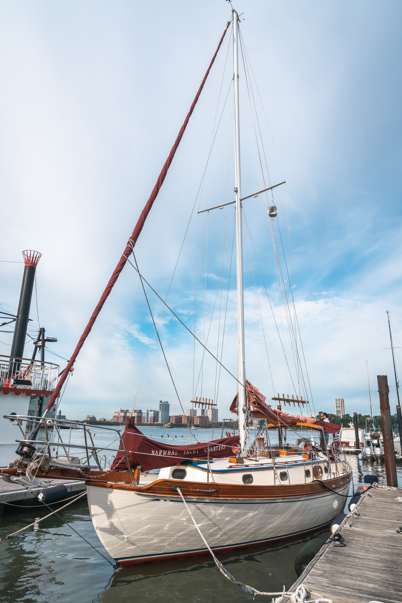 A classic wooden sailboat with red trim and white hull sits moored at a marina dock, its tall mast and rigging silhouetted against a partly cloudy sky with city buildings visible across the water