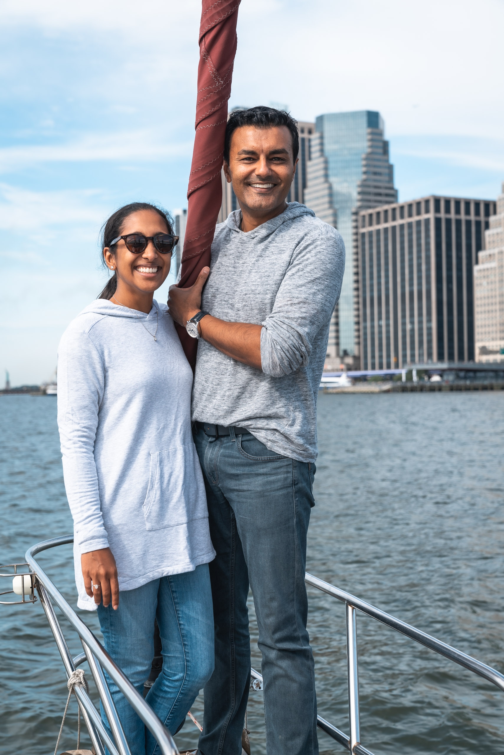 A smiling couple stands together on a boat deck with a red mast, Manhattan's skyline and waterfront buildings visible behind them under a partly cloudy sky