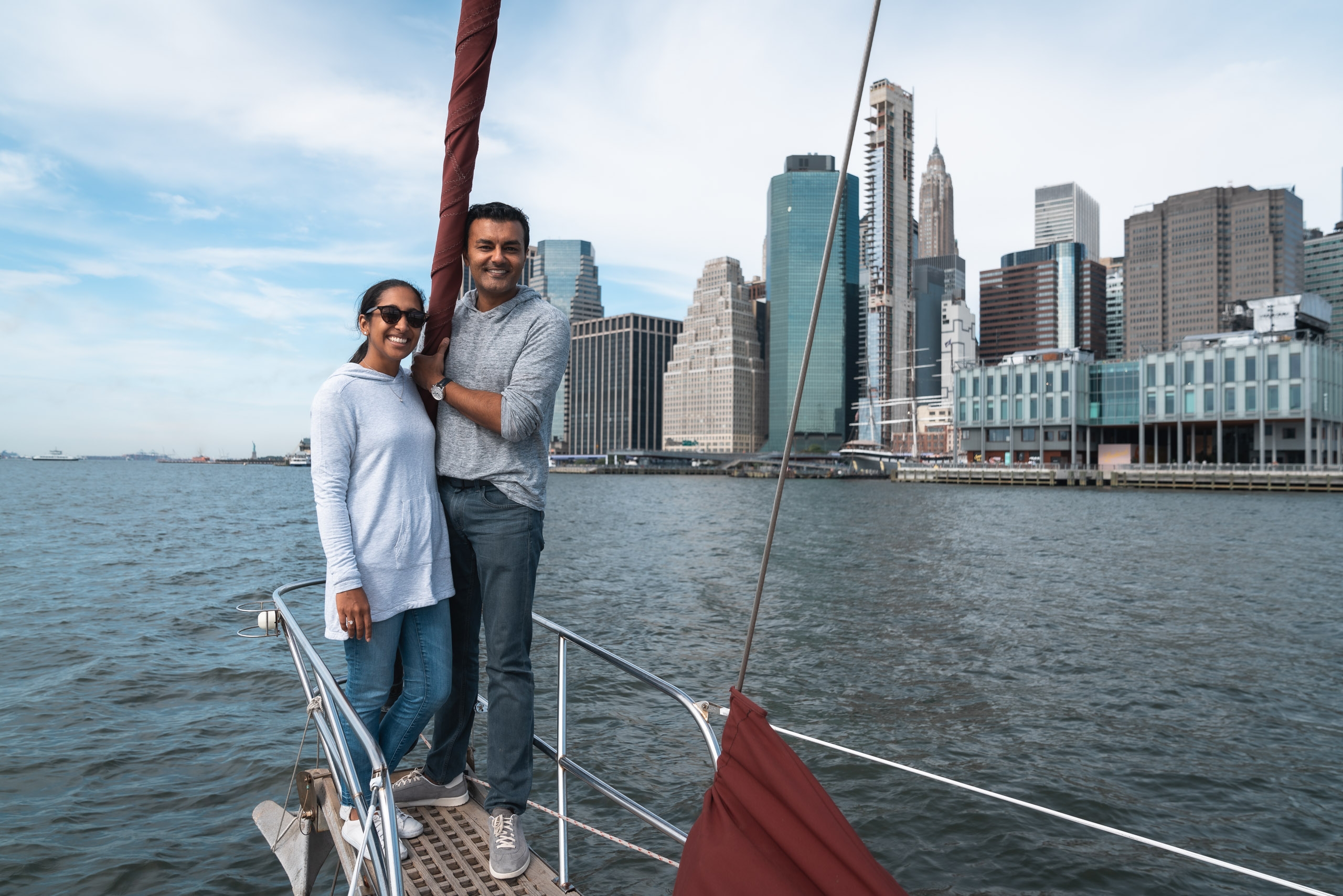 A couple stands together on a sailboat with Manhattan's skyline and tall buildings visible across the blue water behind them under partly cloudy skies