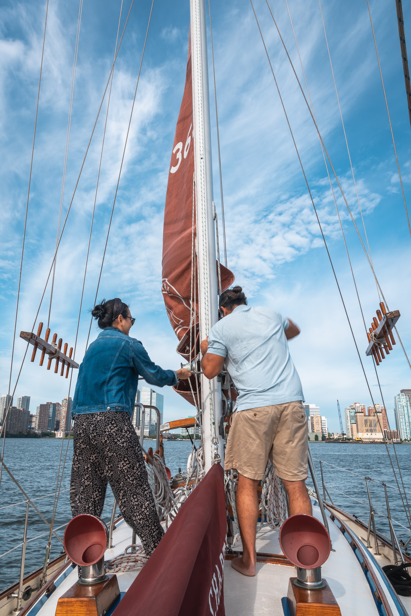 Two crew members adjust a burgundy sail on a sailing vessel with Manhattan's skyline visible across the blue water under a partly cloudy sky