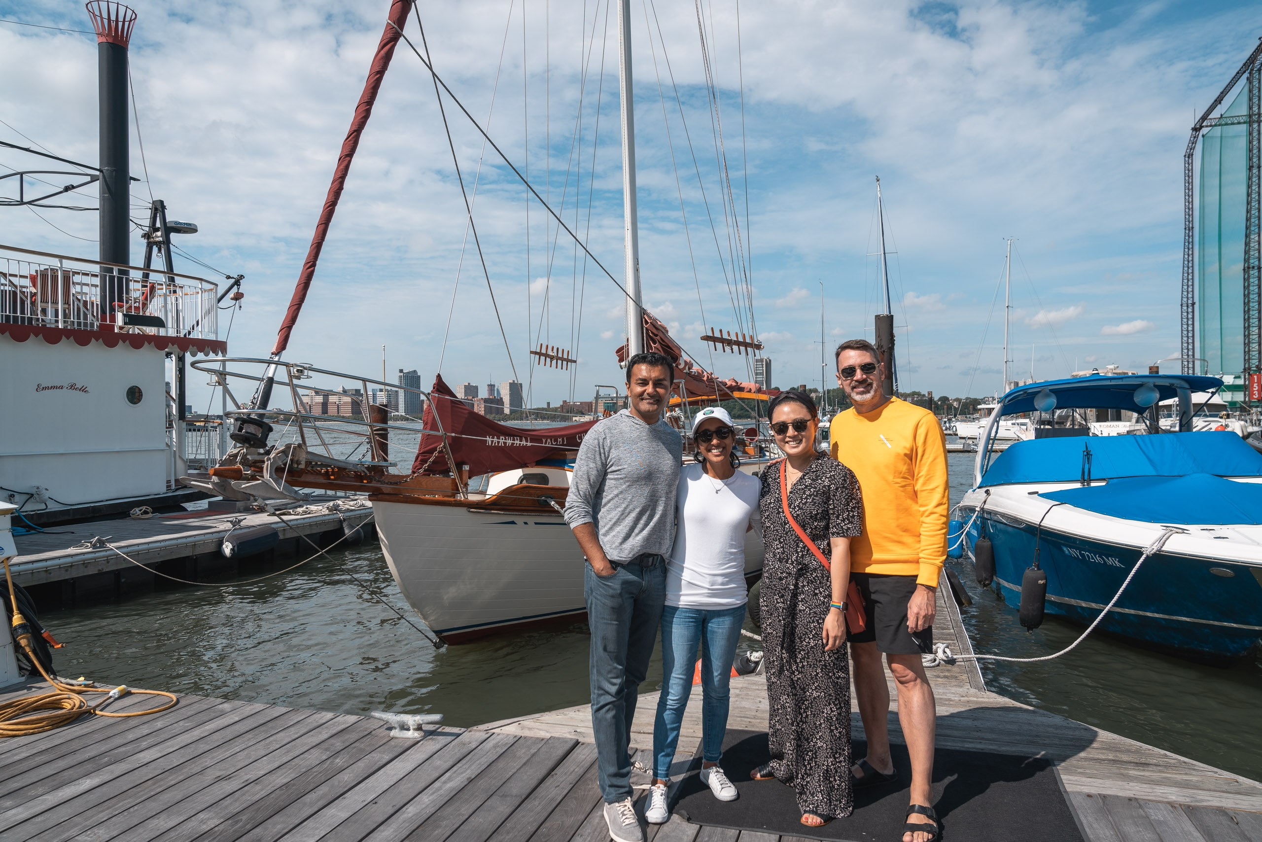 Four people stand on a wooden dock surrounded by moored sailboats and yachts, with a historic sailing ship and city skyline visible under partly cloudy skies