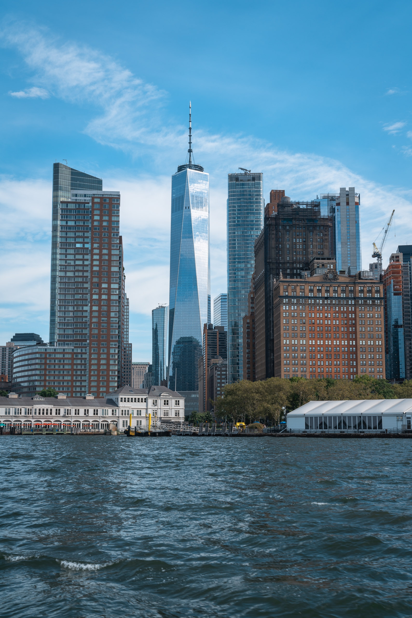 Manhattan's skyline rises prominently above the Hudson River waterfront, featuring One World Trade Center's distinctive white and blue tower amid surrounding residential and commercial buildings under clear blue skies