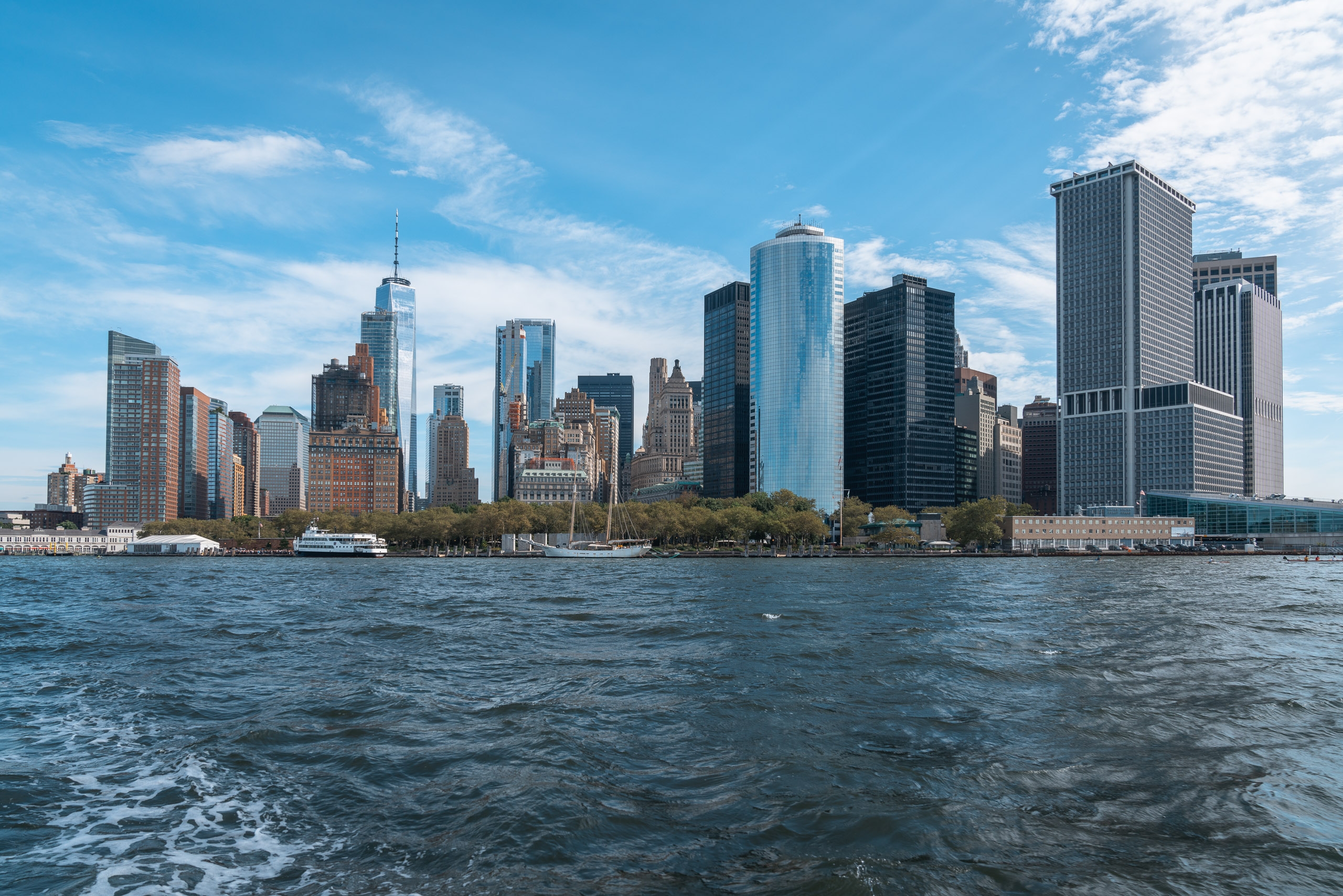 Manhattan's skyline rises dramatically across the East River under clear skies, with diverse architectural styles from historic brownstones to modern glass towers dominating the waterfront view