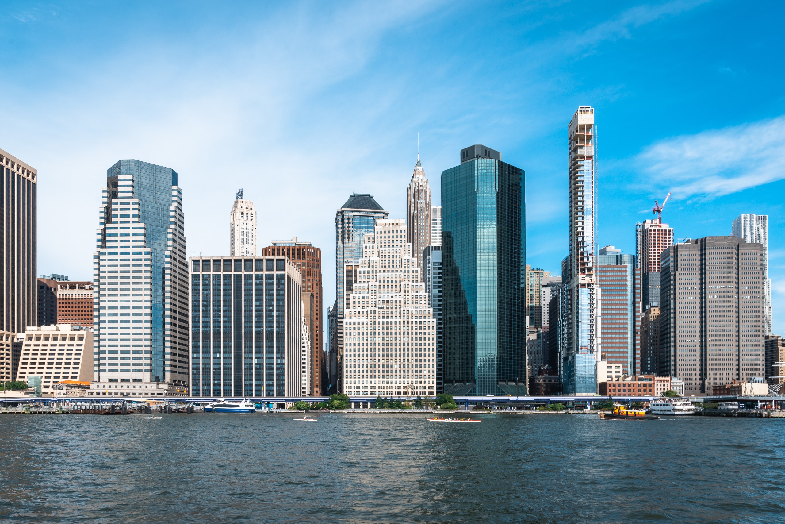 Manhattan's downtown skyline rises dramatically from the East River beneath clear blue skies, with historic and modern towers creating a dense urban profile along the waterfront