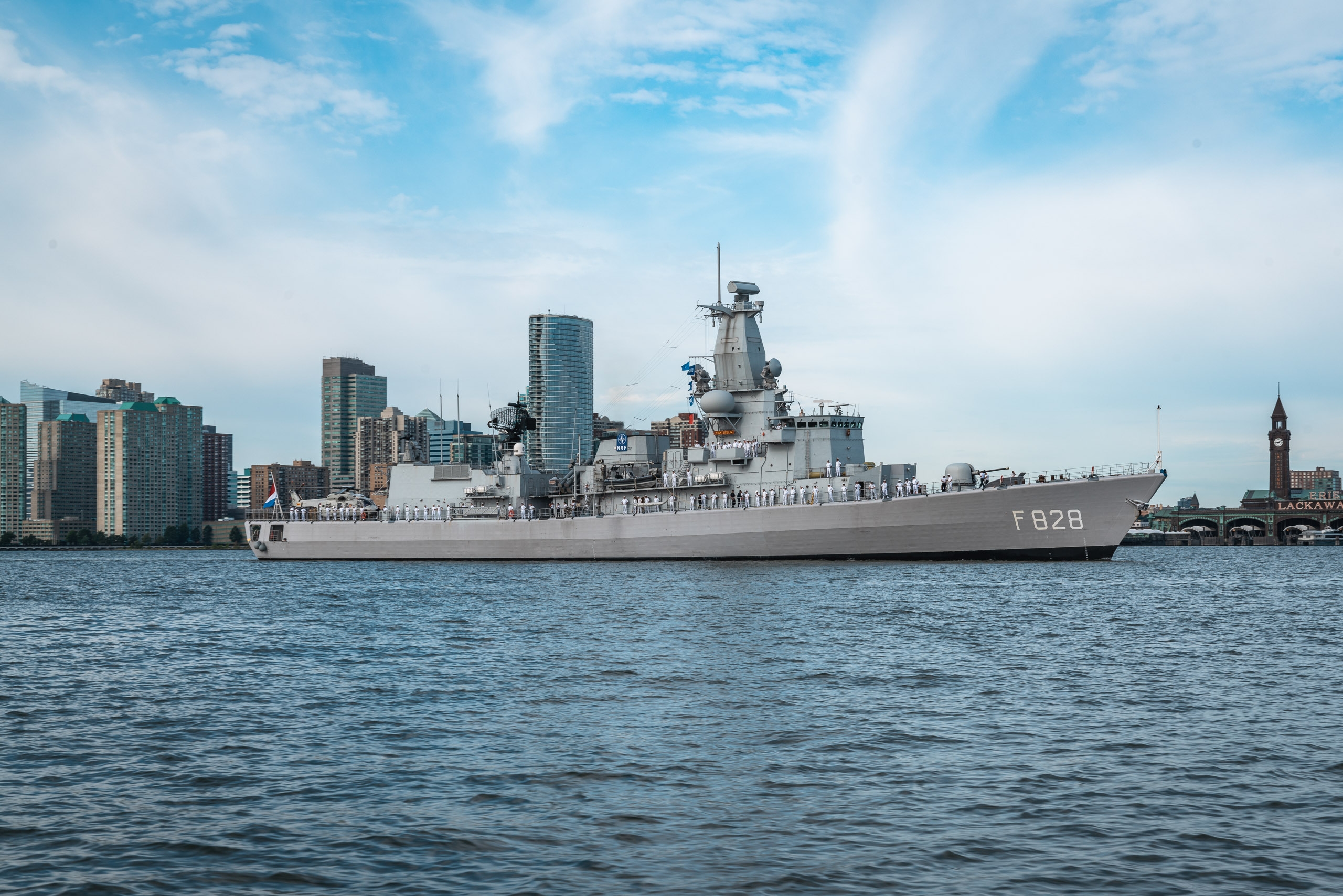 A gray military frigate designated F628 floats on blue-green water with a Manhattan skyline of high-rise buildings and a distant clock tower visible behind it under a partly cloudy sky
