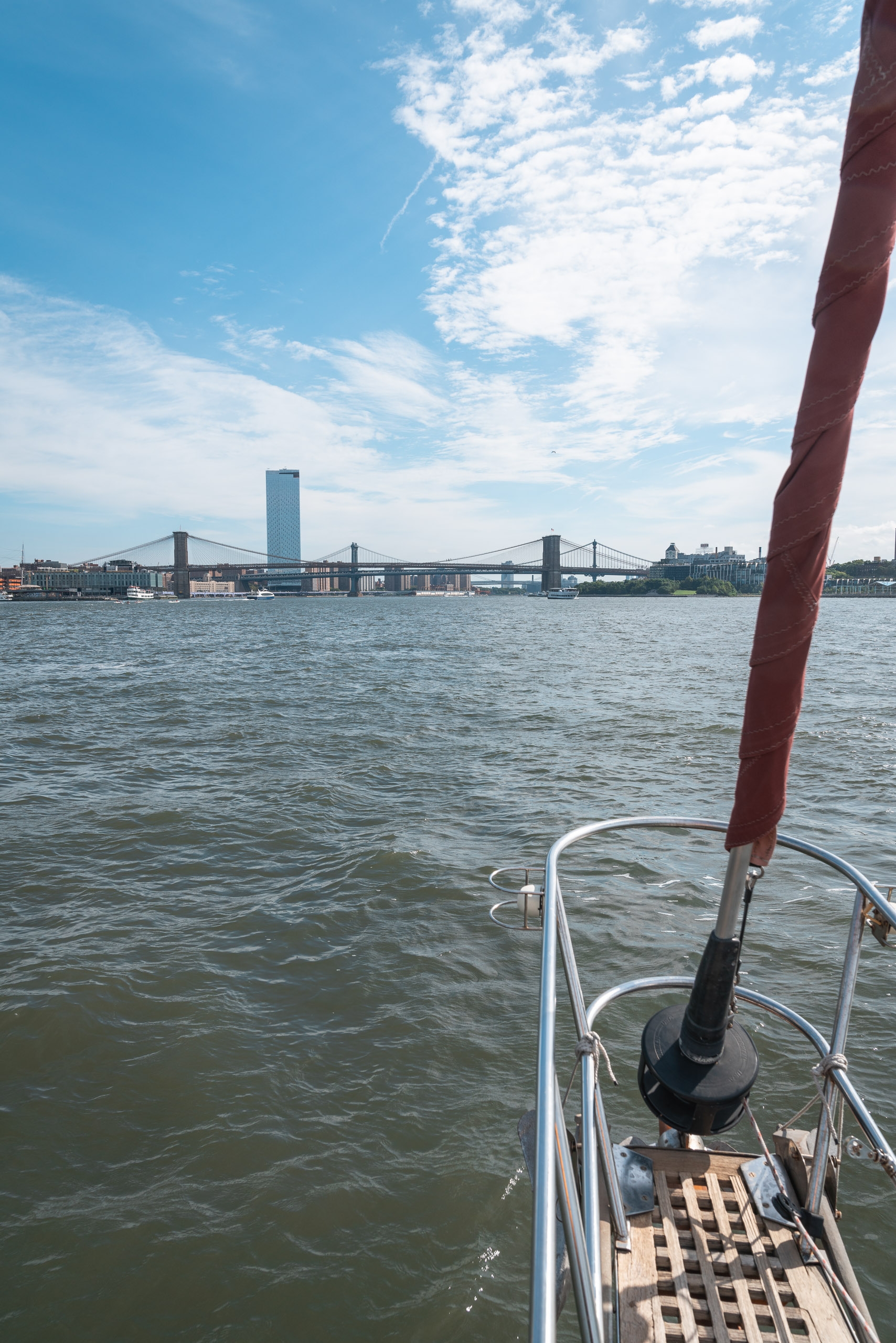 A sailboat's bow with burgundy mast extends toward the Brooklyn Bridge spanning the East River under a partly cloudy blue sky, with Manhattan's waterfront visible in the distance