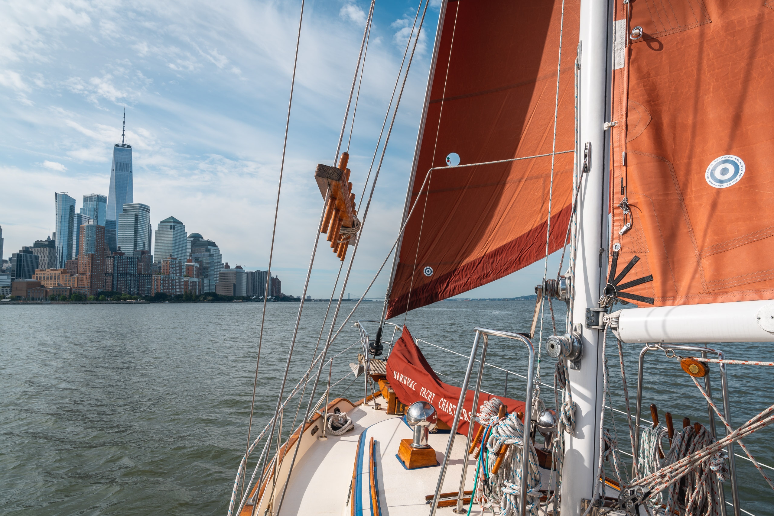 A sailboat with rust-colored sails glides across calm water toward the Manhattan skyline, captured from the deck with rigging and hardware in sharp detail