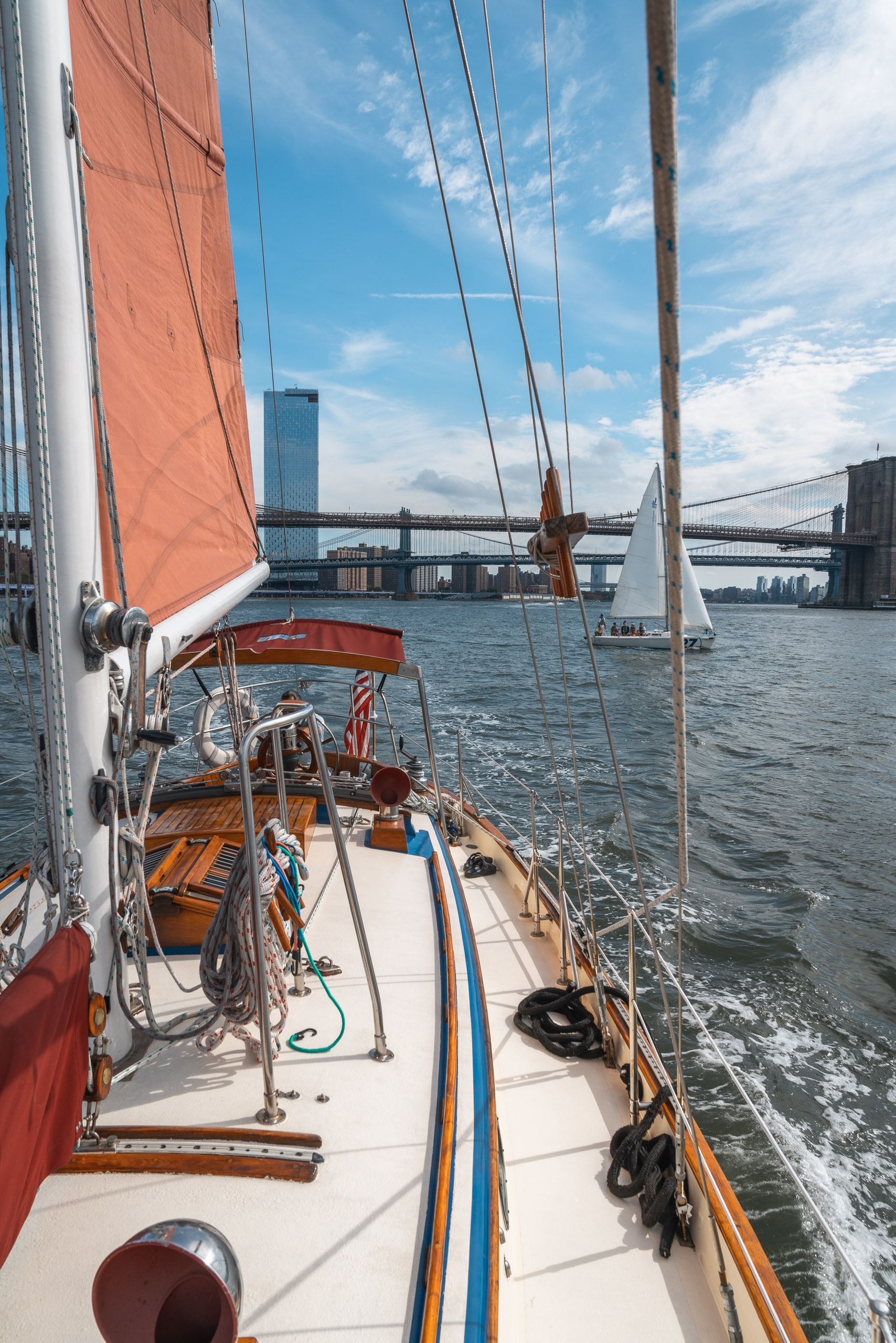 A wooden sailboat with rust-red canvas sails glides past the Brooklyn Bridge on sparkling blue water under partly cloudy skies, with Manhattan's skyline visible in the distance