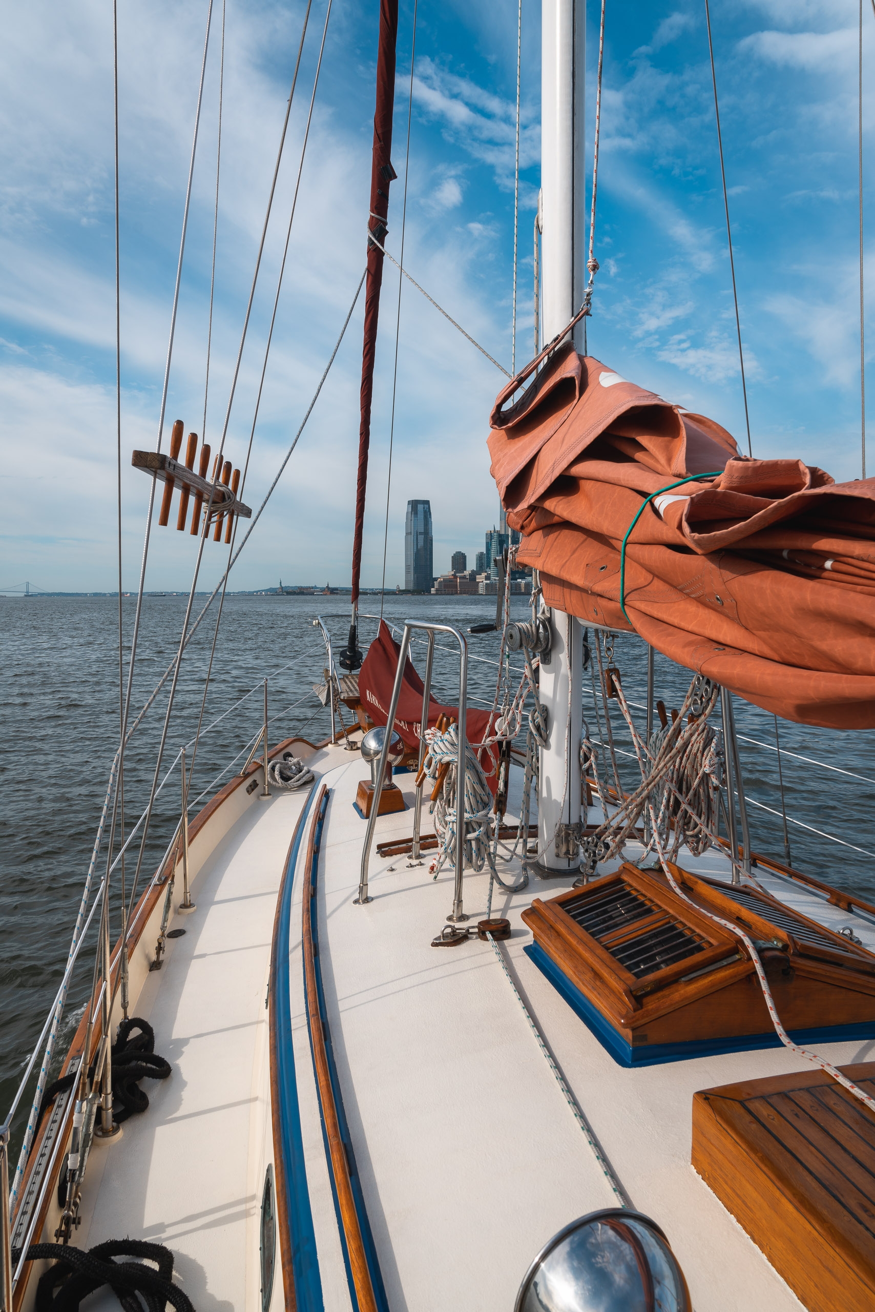 Sailboat deck stretches toward a distant urban skyline with furled orange sails, taut rigging, and wooden details under bright blue sky and scattered clouds