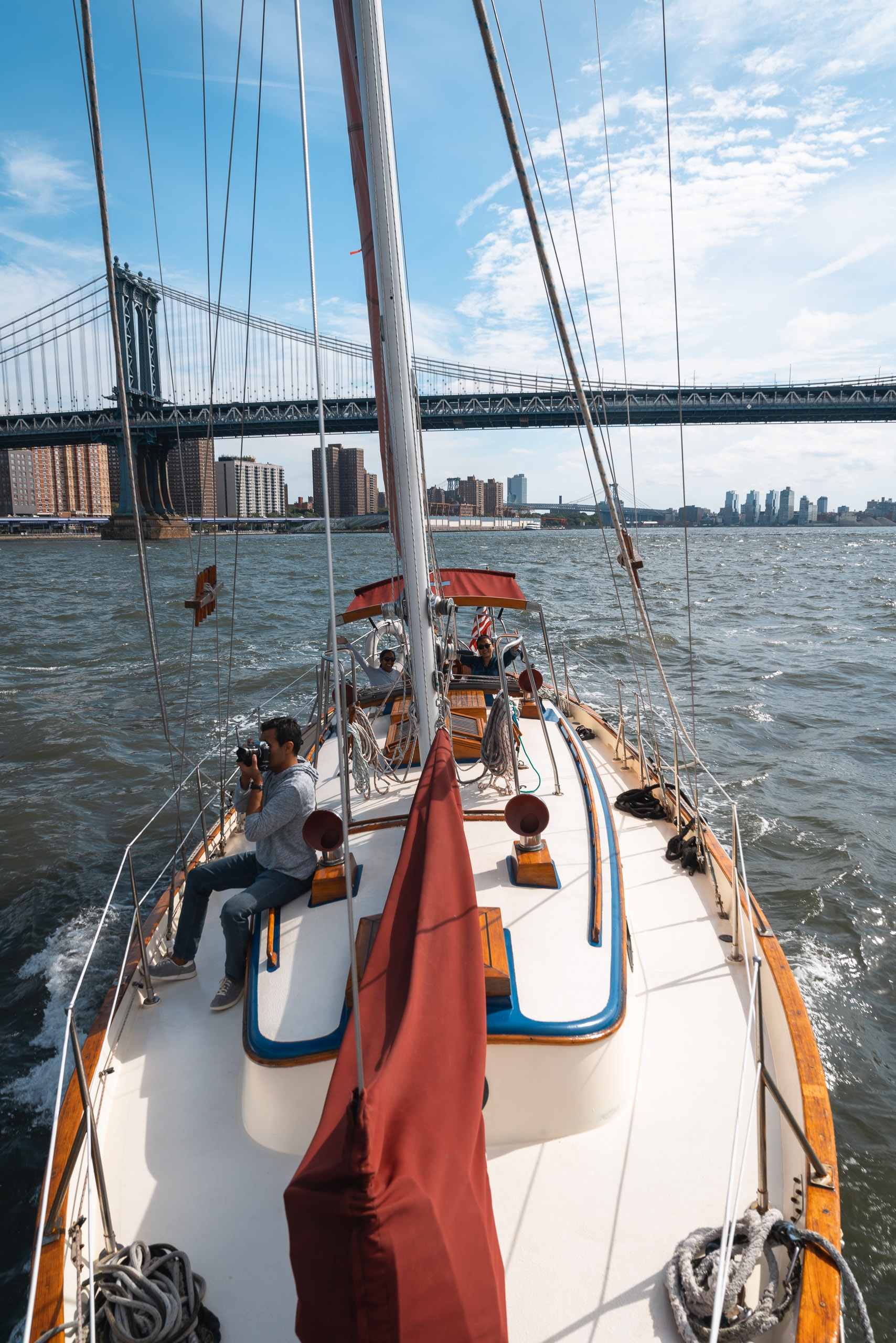 A sailboat with passengers glides across the East River under the Brooklyn Bridge, with Manhattan's skyline and distant Brooklyn shoreline visible beneath a partly cloudy sky