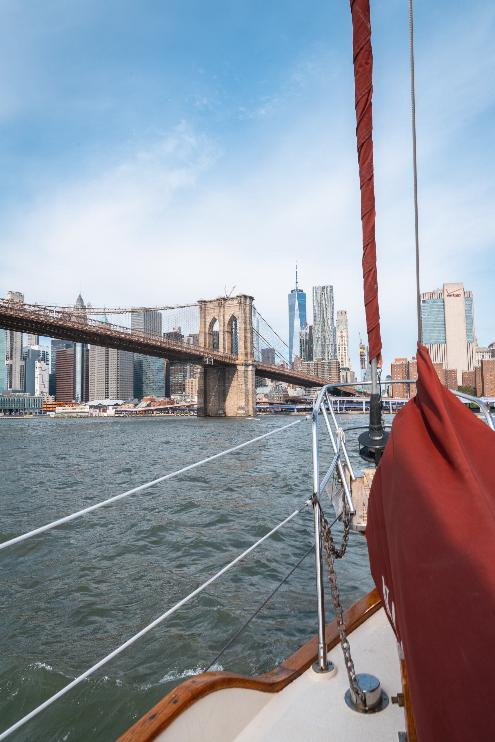 A sailboat's red canvas and wooden rail frame the view toward Manhattan's skyline and the Brooklyn Bridge spanning the East River under clear blue skies